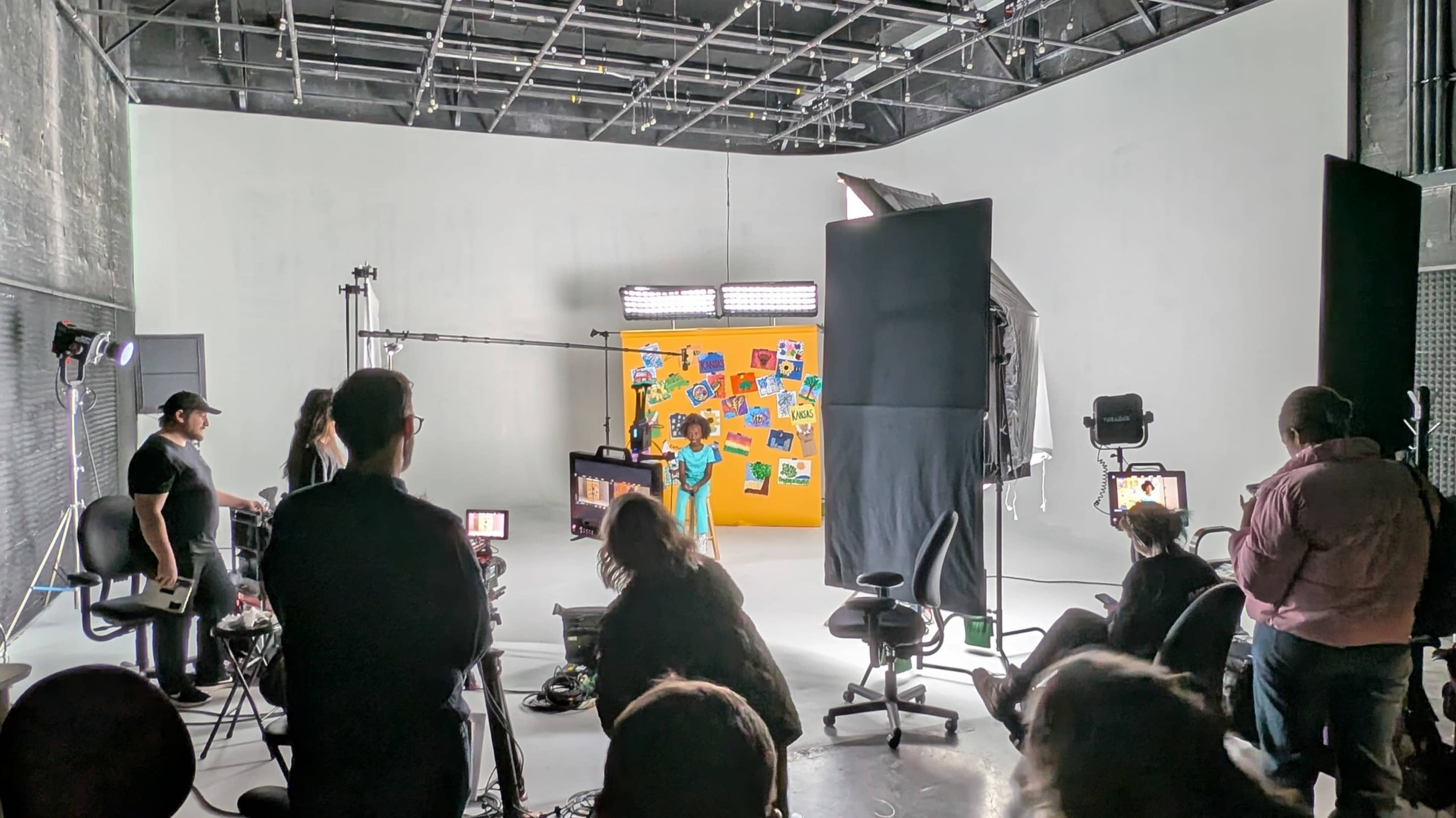 A group of people work in a studio setting with lighting equipment and a colorful backdrop, while a performer engages with a camera.