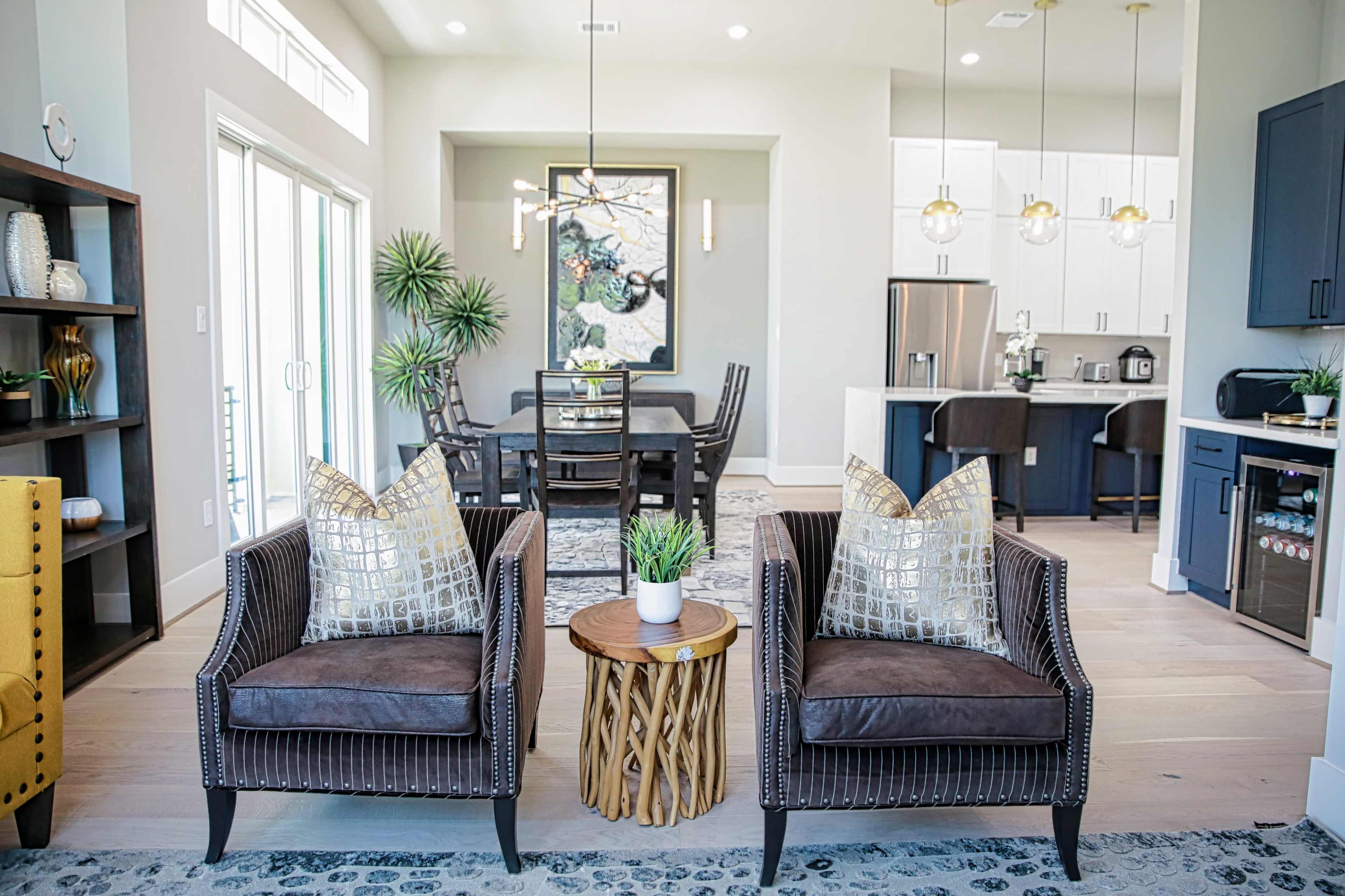 The image shows a modern living area with two upholstered chairs facing a small wooden table, showcasing a dining area and kitchen in the background.