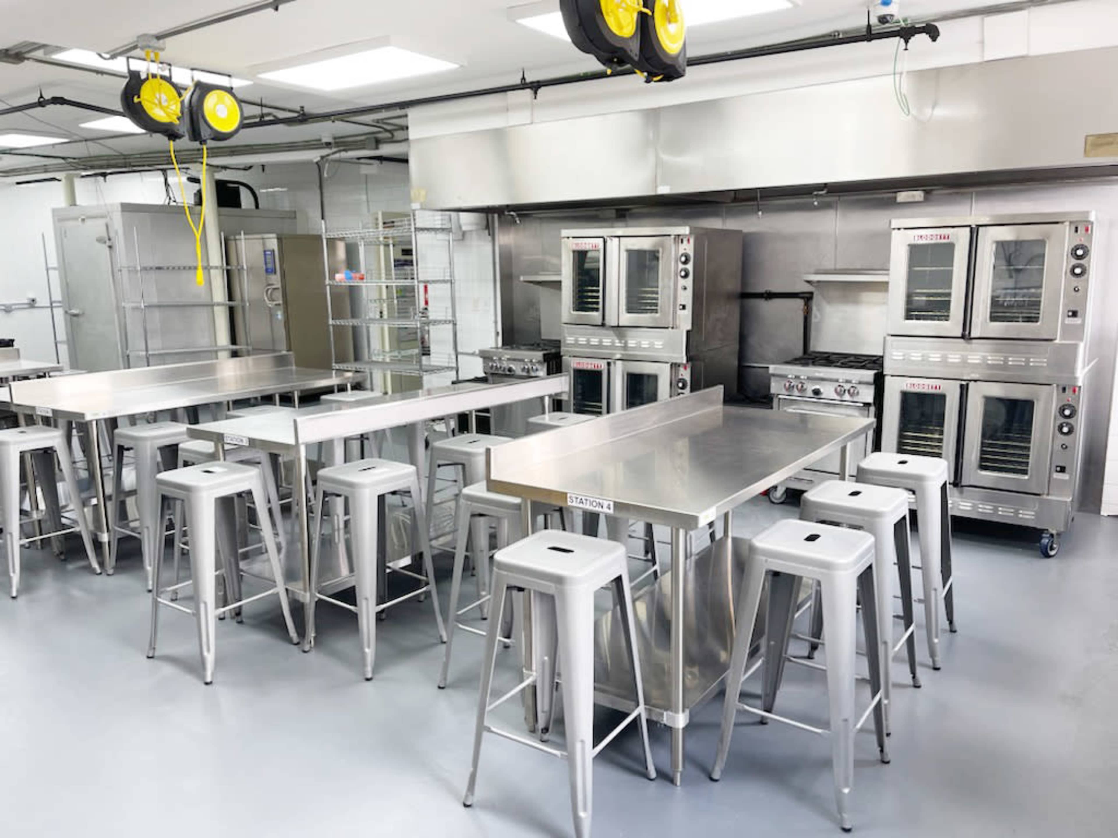 A commercial kitchen is equipped with stainless steel tables, high stools, and multiple ovens and cooking appliances.