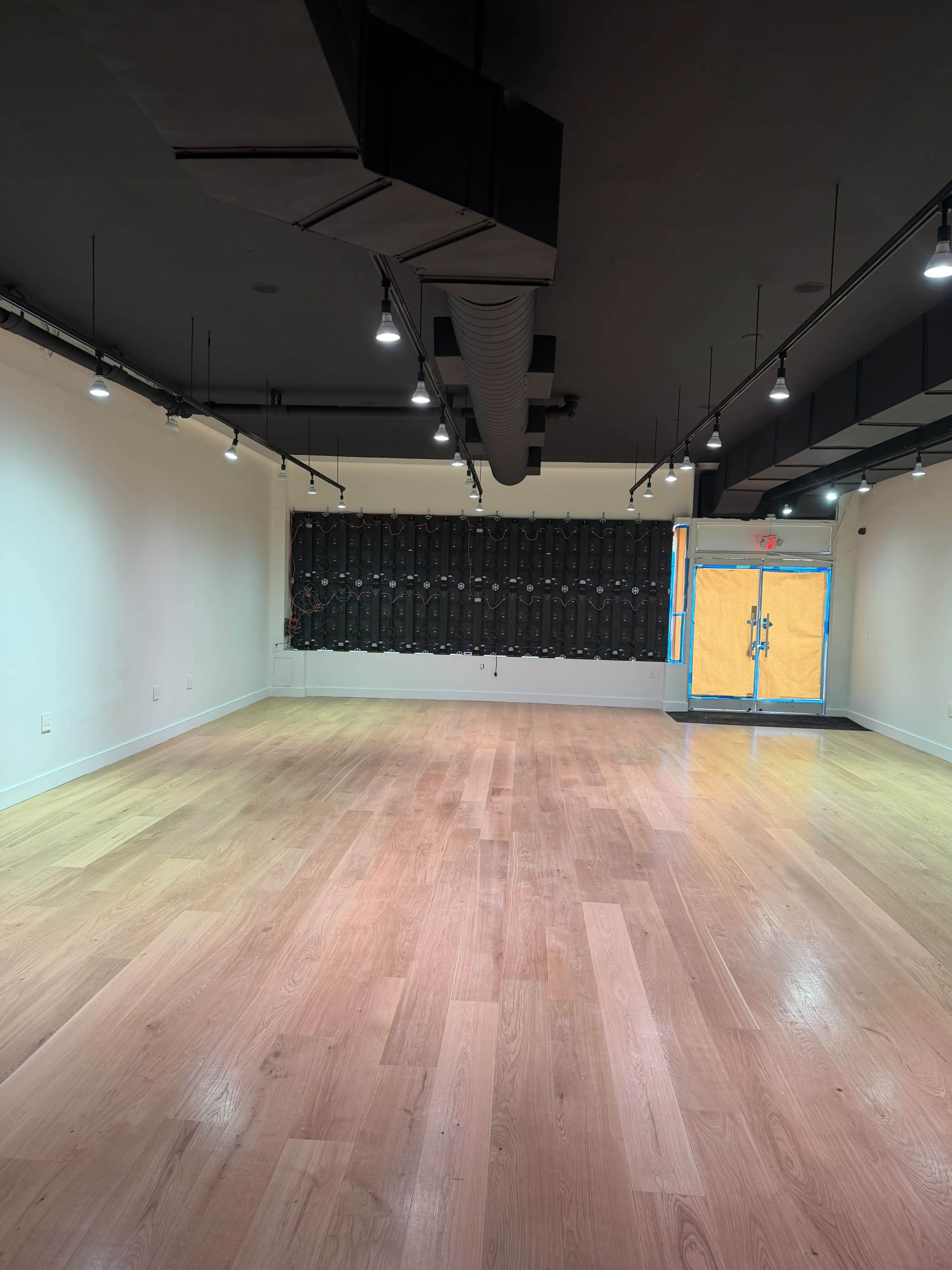 The image shows an empty room with a wooden floor, white walls, and a dark wall featuring a grid pattern at one end, along with a double door painted yellow.