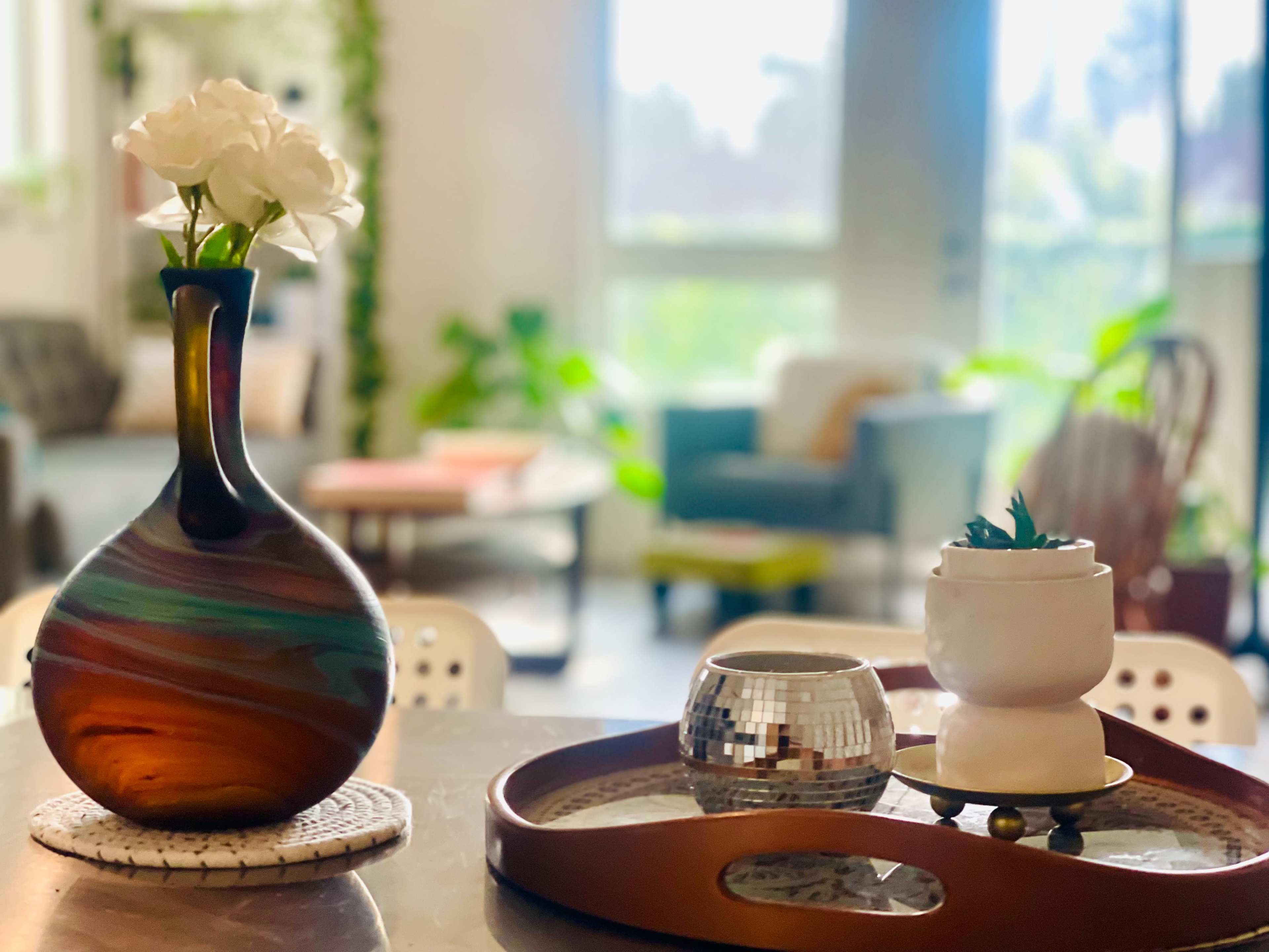 The image shows a colorful vase with flowers next to a disco ball and a small potted plant on a wooden tray, with a bright, airy living room in the background.