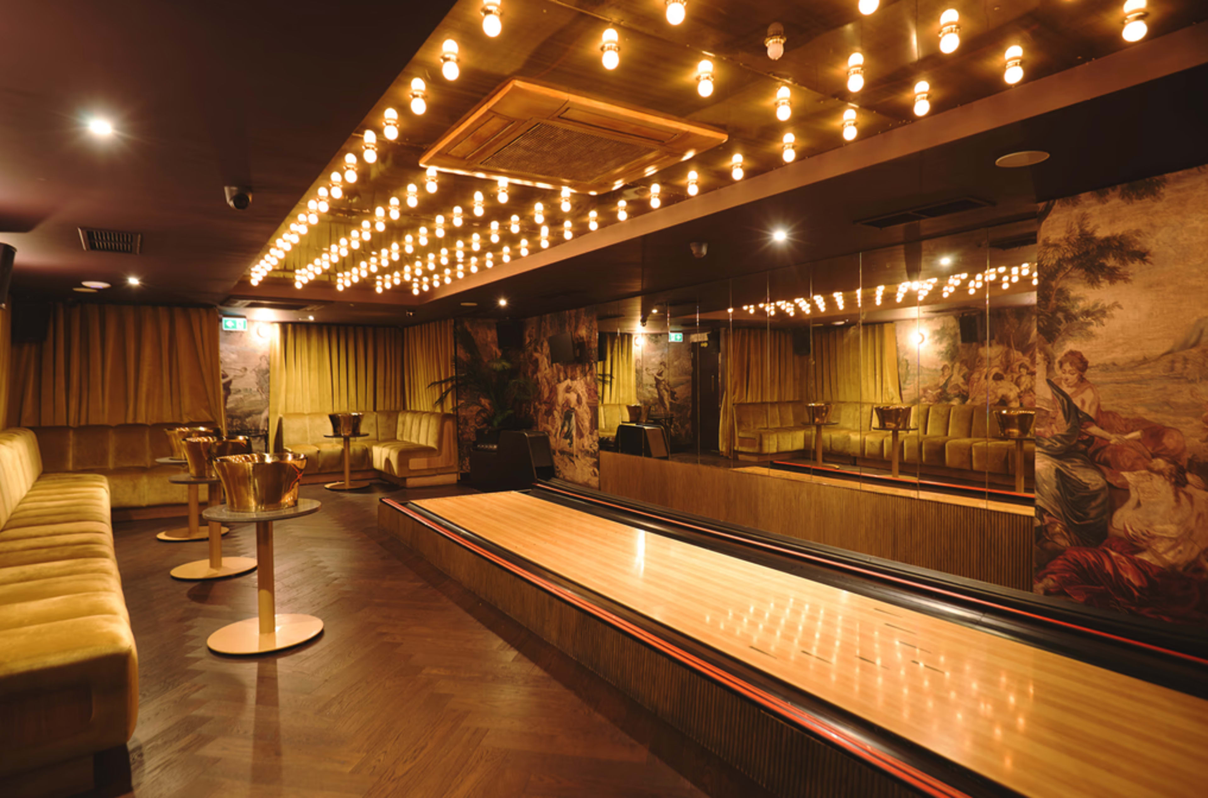 The image depicts a stylish bowling alley with a polished wooden lane, plush seating, and decorative lighting, set against ornate wall murals.