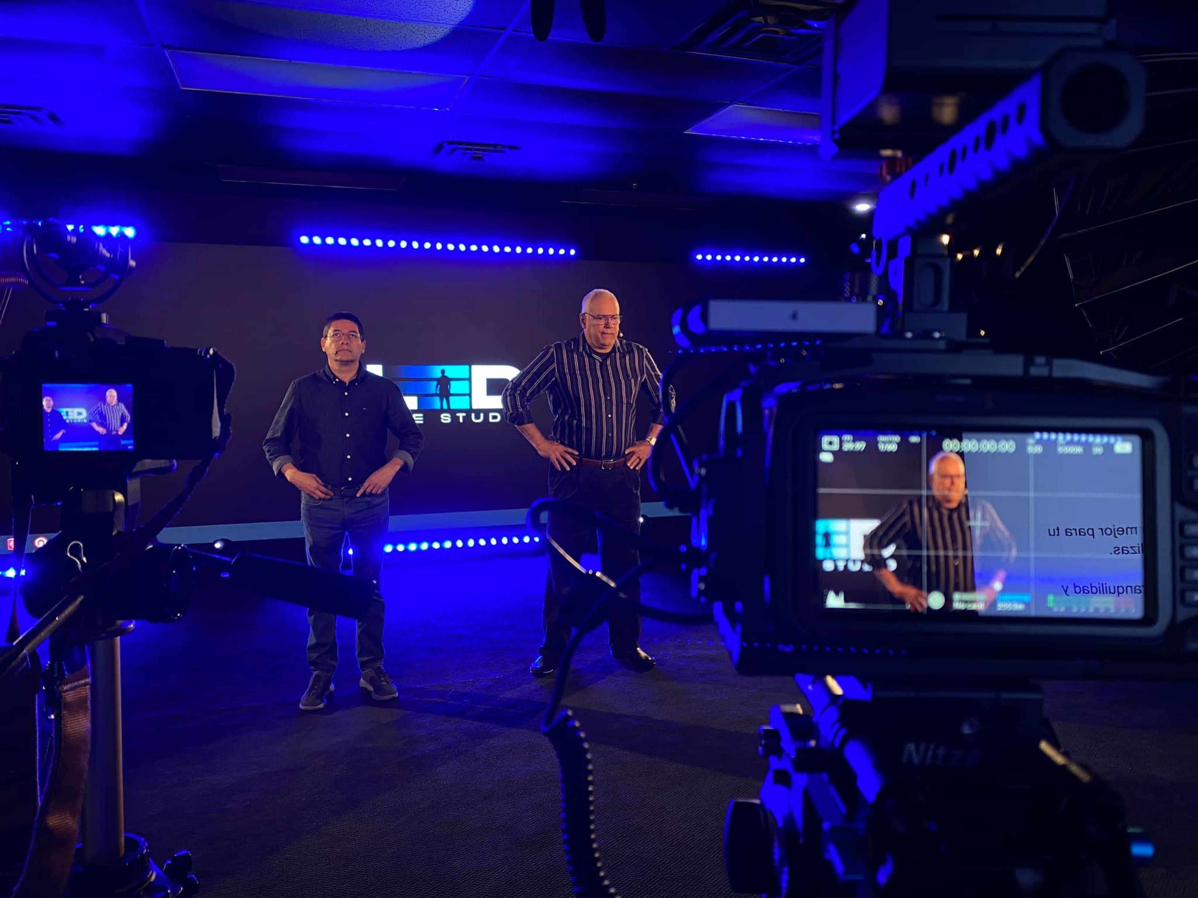 Two men stand in a studio space with blue lighting while cameras are set up to record.