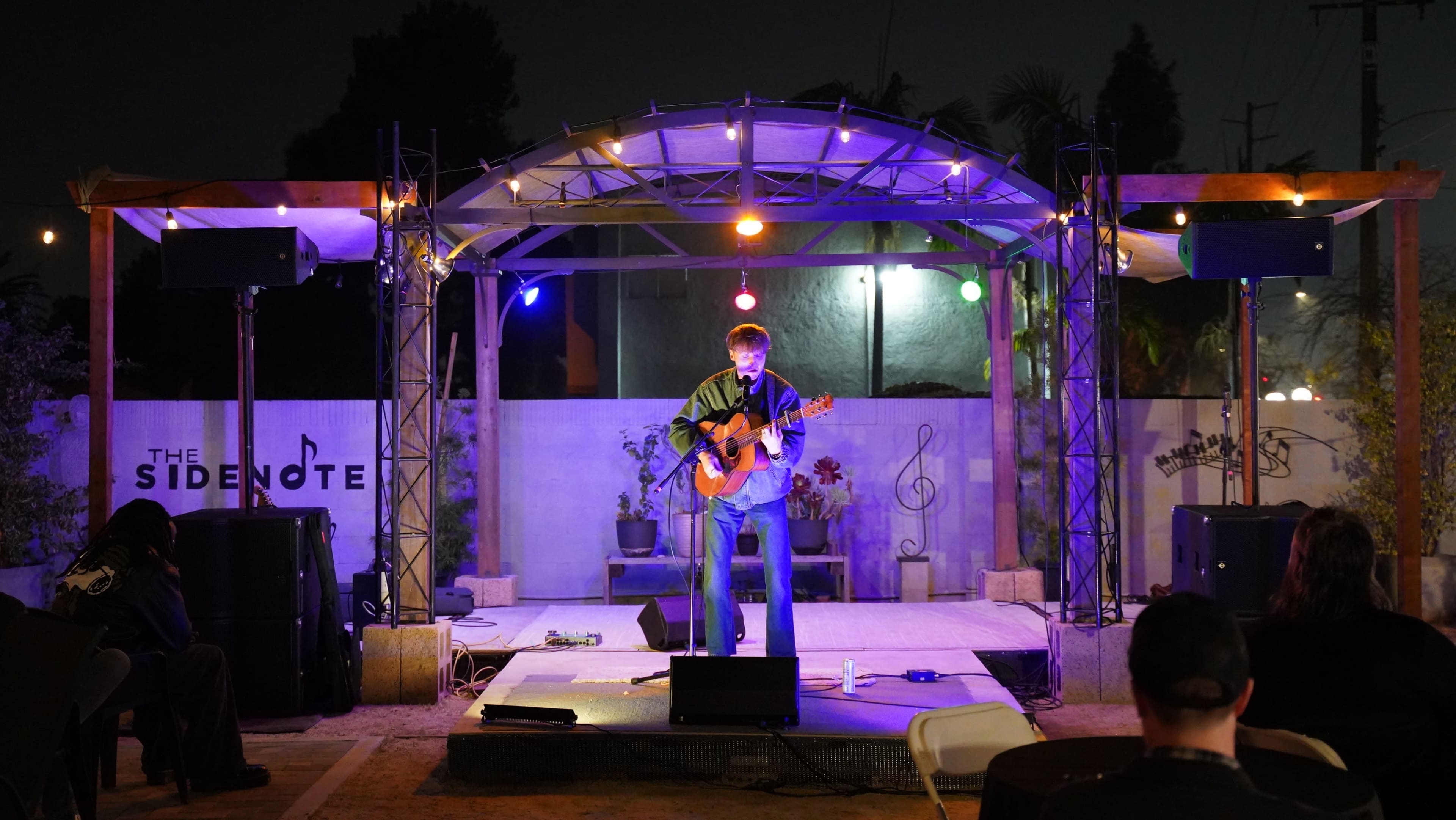 A musician performs on stage with an acoustic guitar under colorful lights at an outdoor venue called The Sidenote.