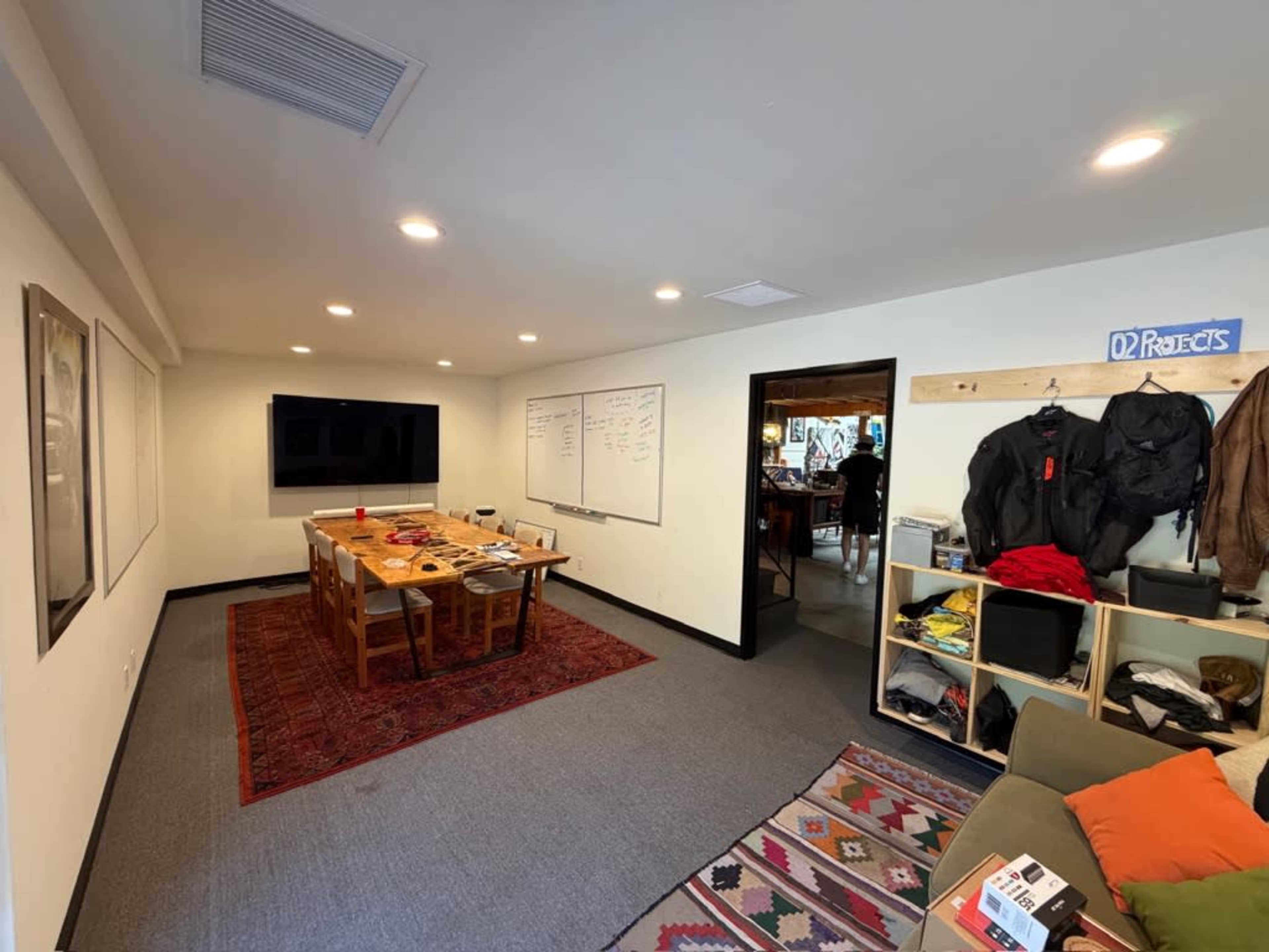 The image shows a modern meeting room with a large table, a television on the wall, whiteboards, and shelves filled with various items.