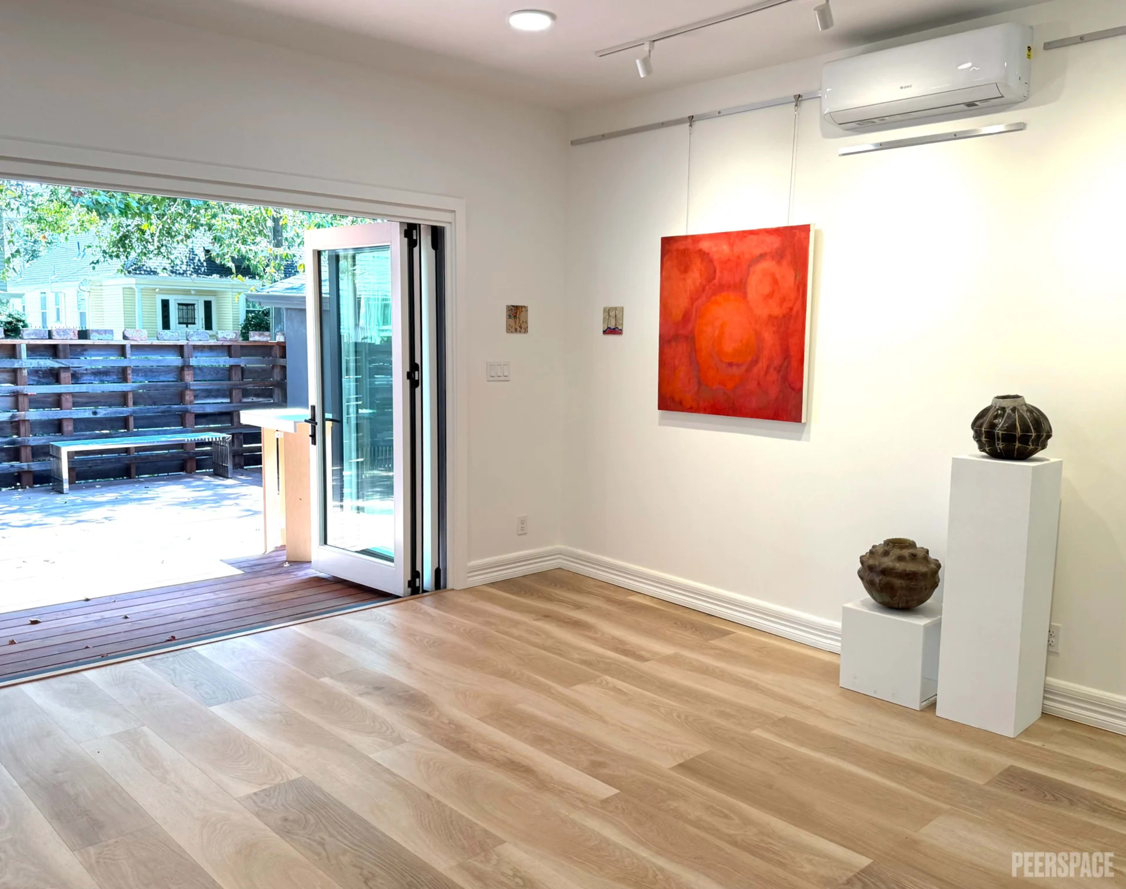 The image shows a spacious, well-lit art gallery featuring a red abstract painting on the wall, with a doorway leading to an outdoor area and two decorative pottery pieces on display.