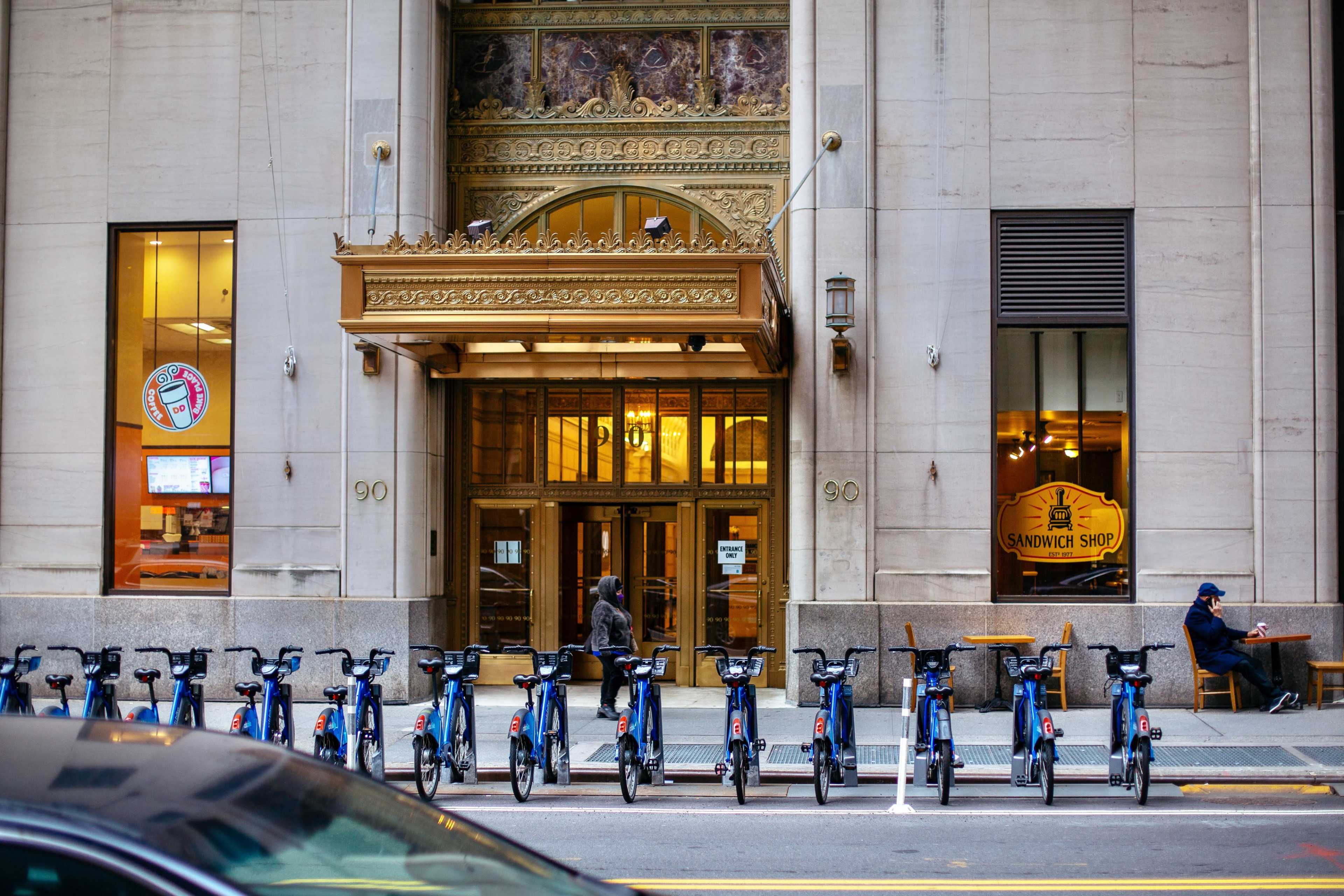 A row of blue bicycles is lined up on a sidewalk in front of a historic building featuring ornate architectural details and a sandwich shop.