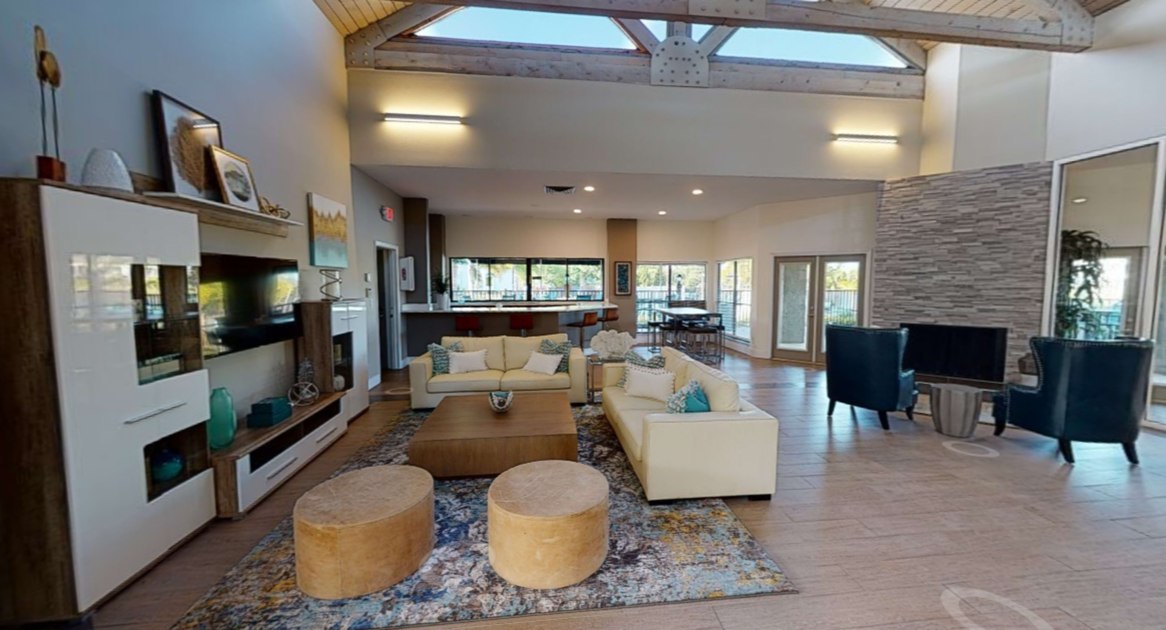 The image shows a modern living room with a high ceiling, light-colored furniture, a large area rug, and large windows letting in natural light.