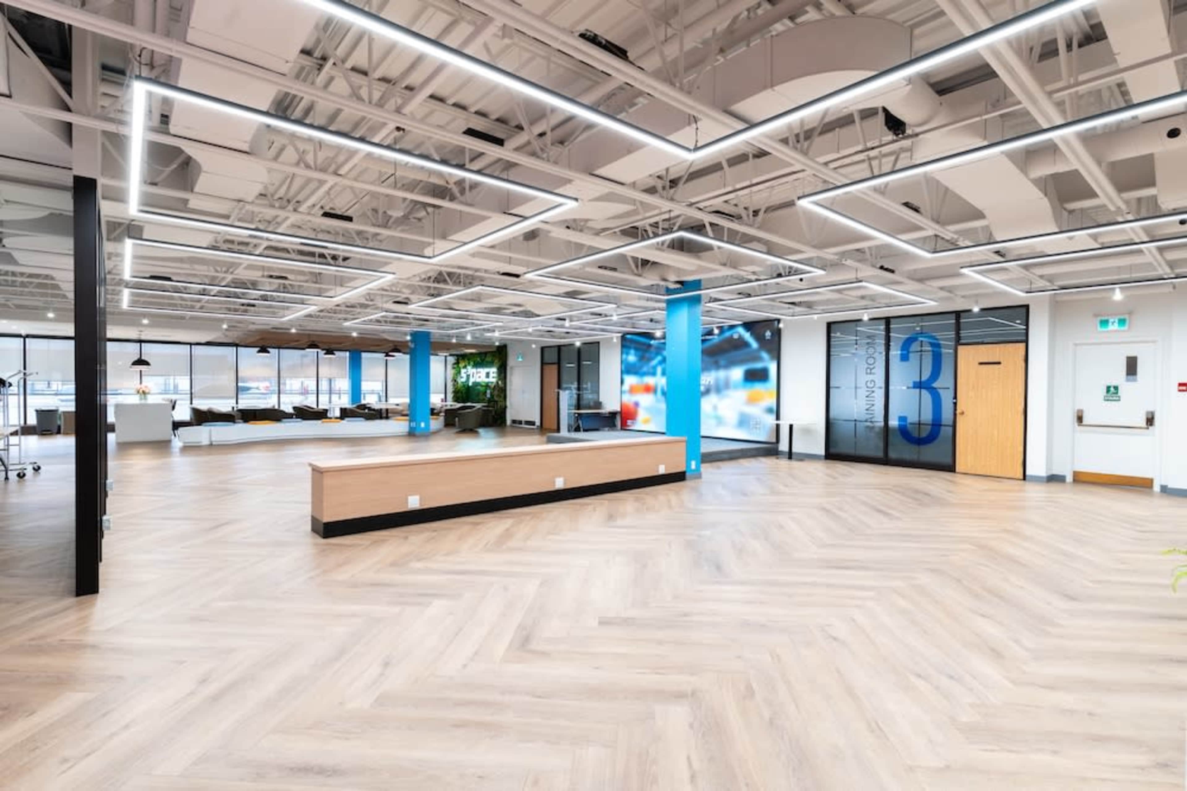 The image shows a modern office space with a large open area, featuring light wooden flooring, a reception desk, and several meeting rooms with glass partitions.