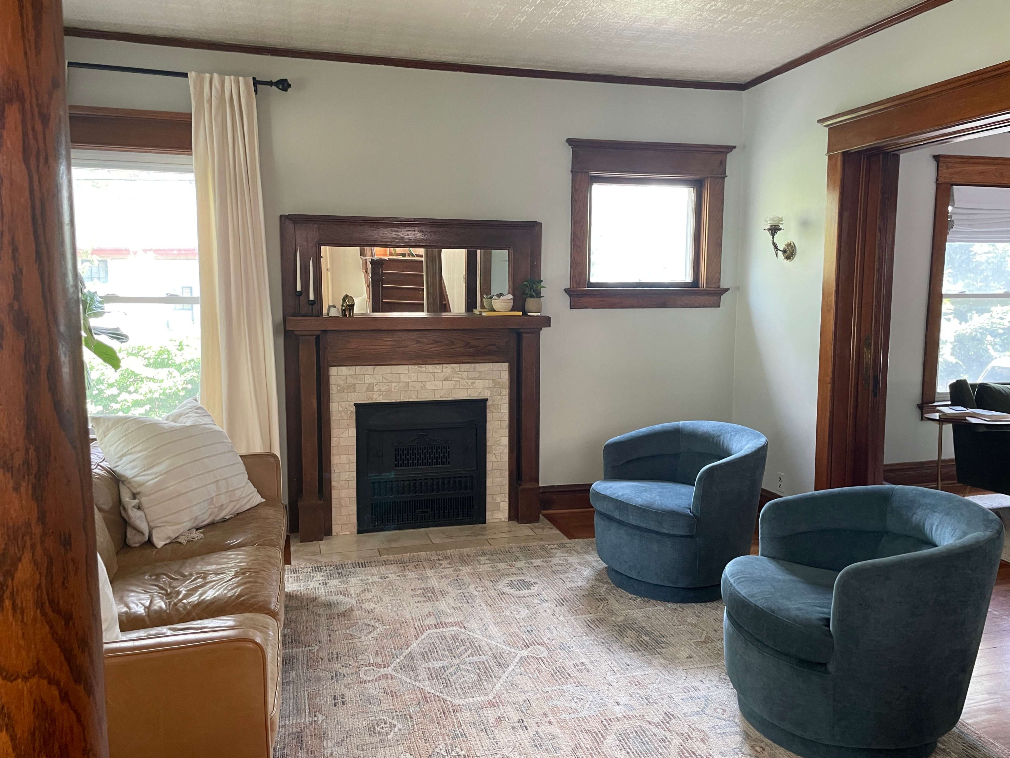 The image shows a cozy living room with a tan leather sofa, two blue chairs, a fireplace with a stone mantle, and large windows allowing natural light to enter.