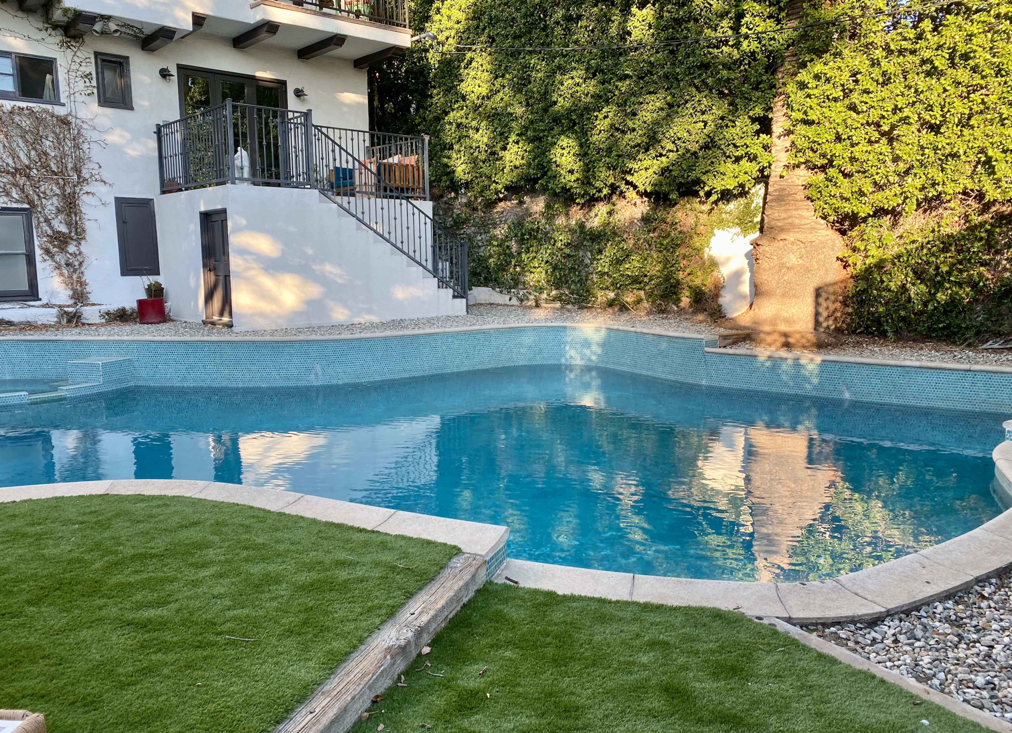 A backyard swimming pool surrounded by a stone patio and lush greenery.