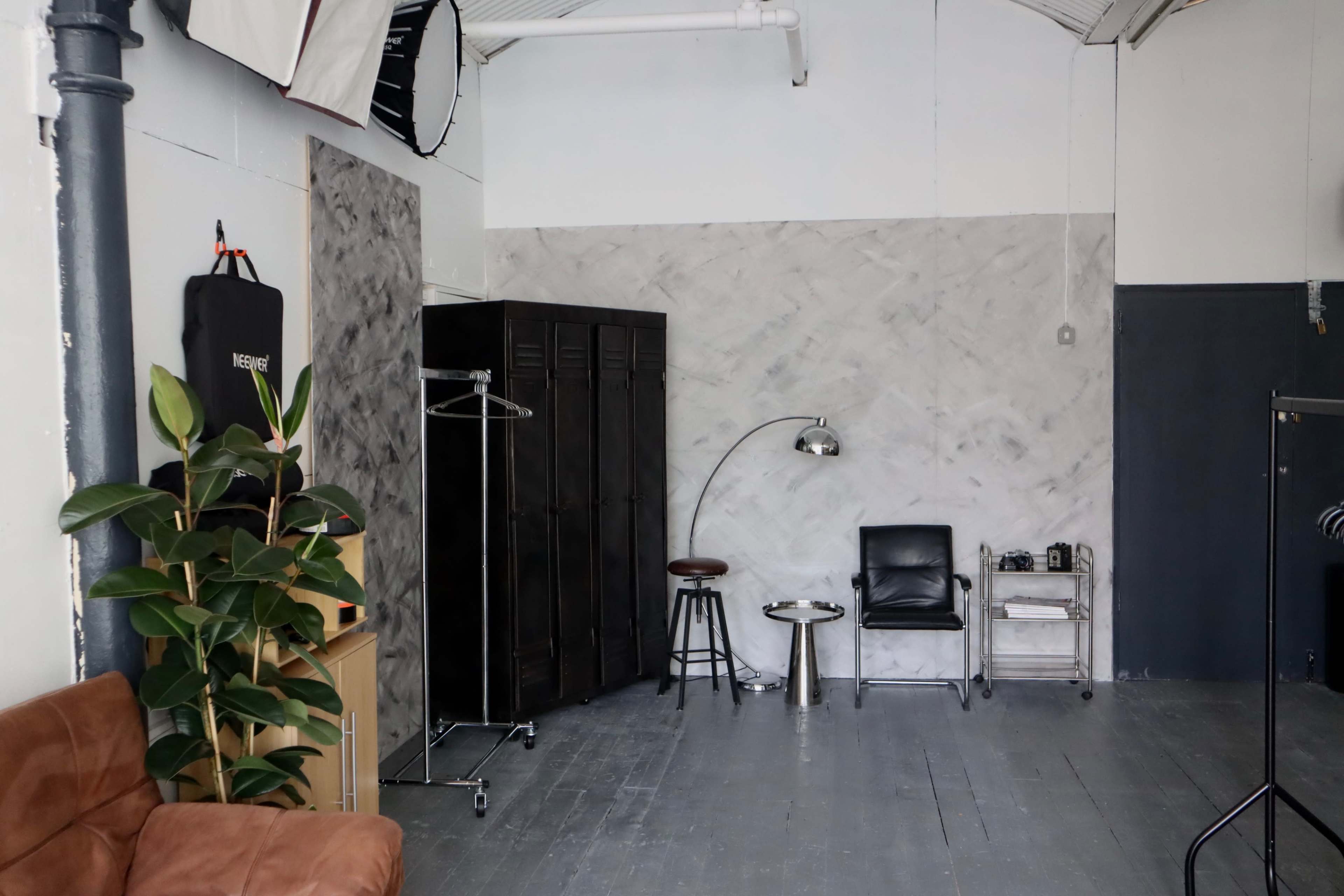 The image depicts a minimalist studio space featuring a brown leather couch, a black wardrobe, a metal stool, a floor lamp, and a clothing rack against a light-colored wall.