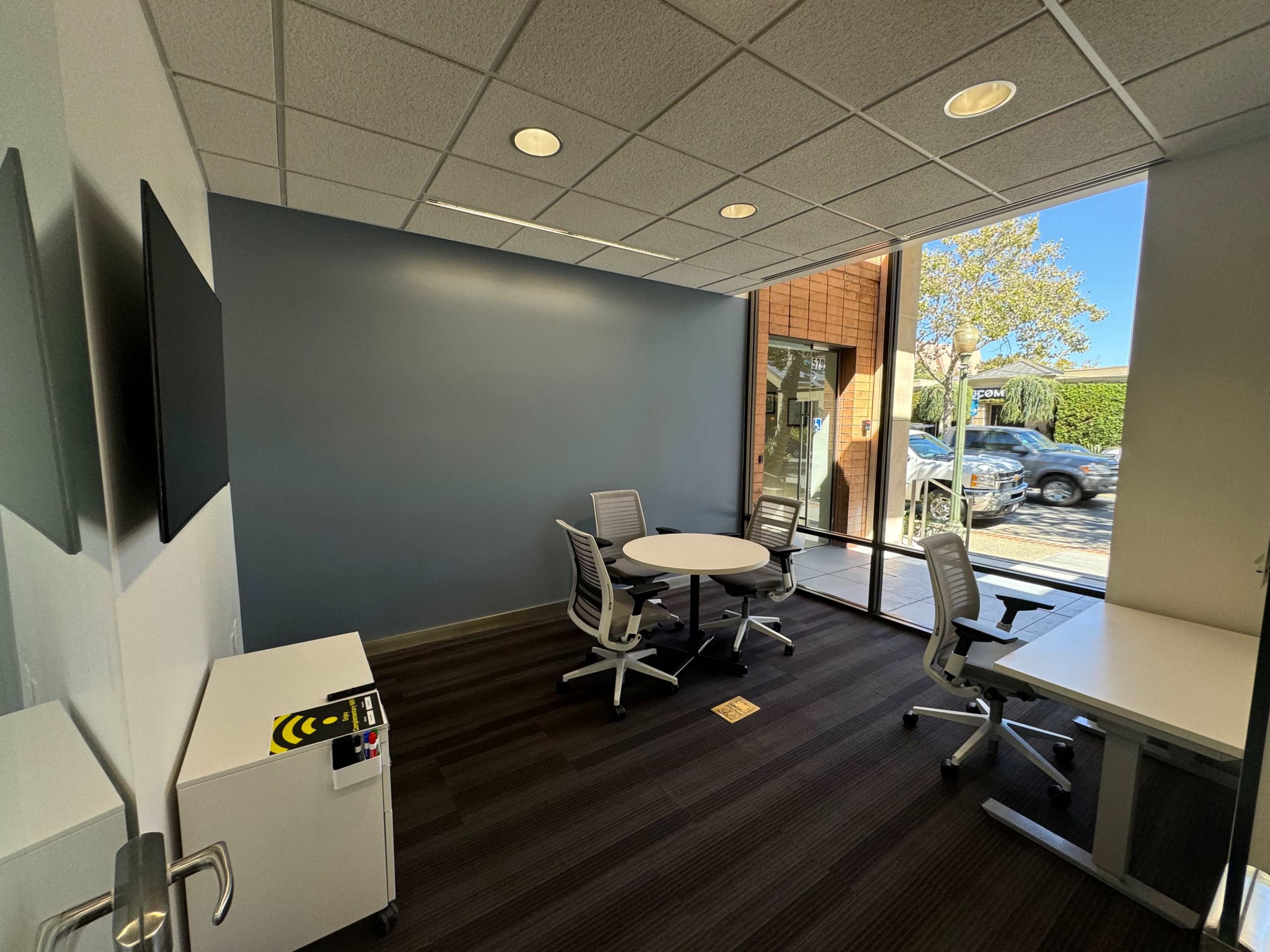 A modern office meeting room with a round table and four chairs, large windows, and a white desk.