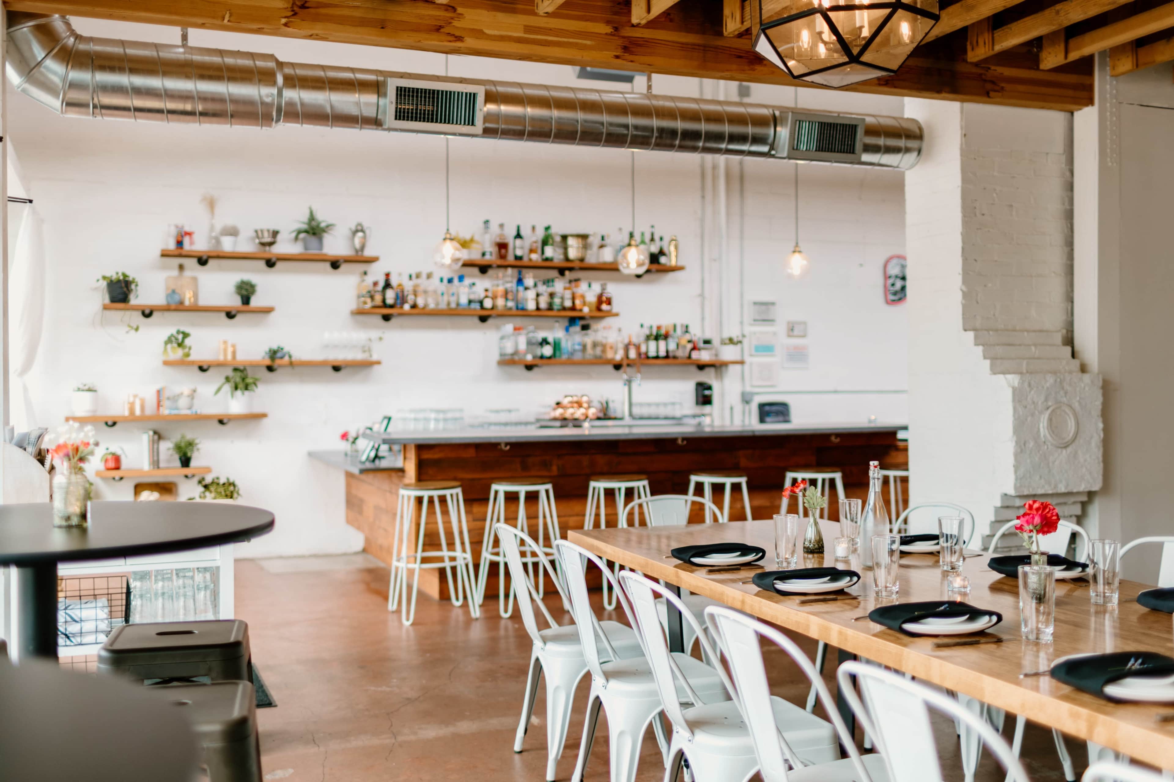 The image shows a modern restaurant interior with wooden shelves displaying various plants and bottles, a long wooden communal table with white chairs, and a bar area in the background.
