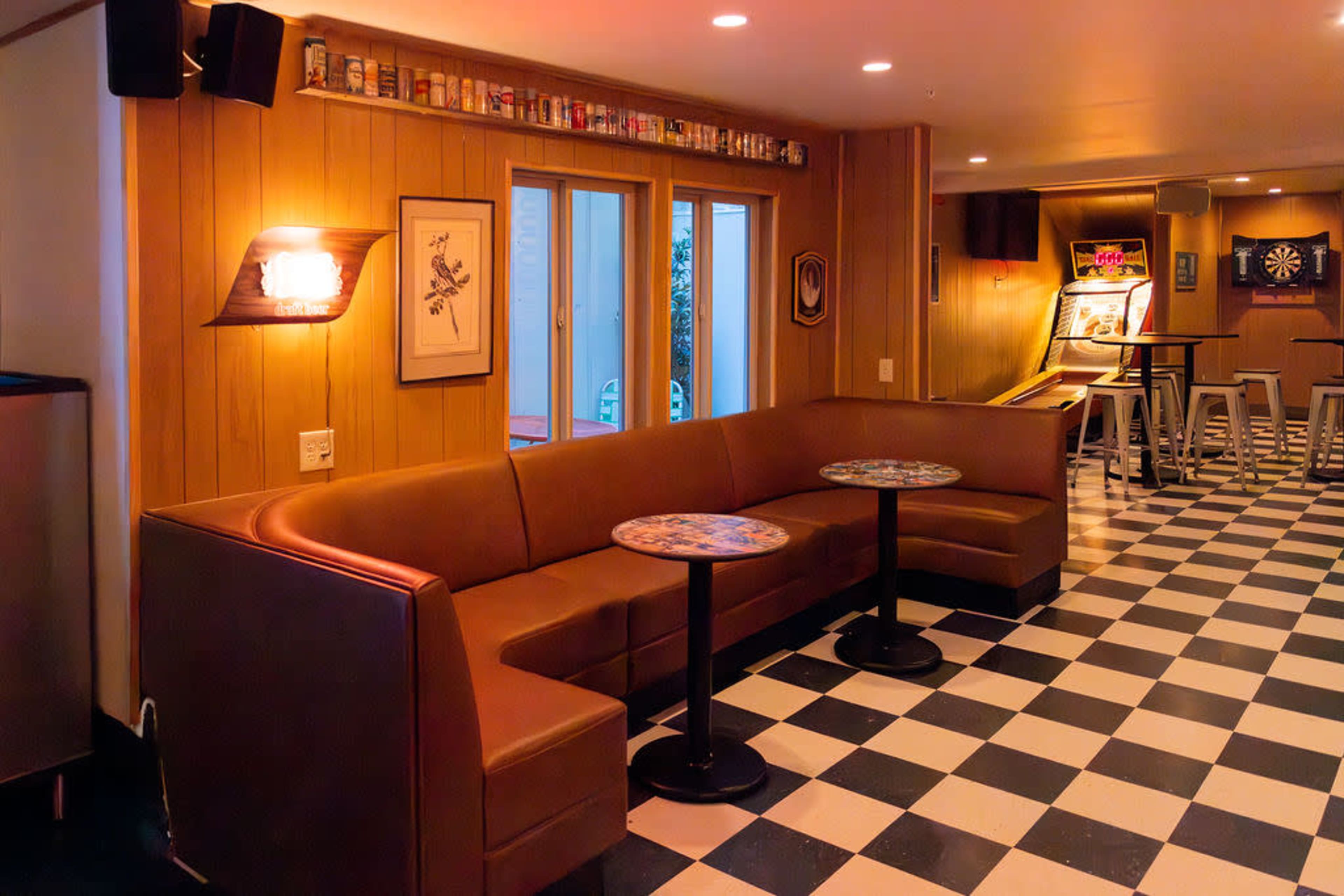 The image shows a cozy lounge area with a brown sectional couch, circular tables, and checkered flooring, featuring a bar area and game machines in the background.