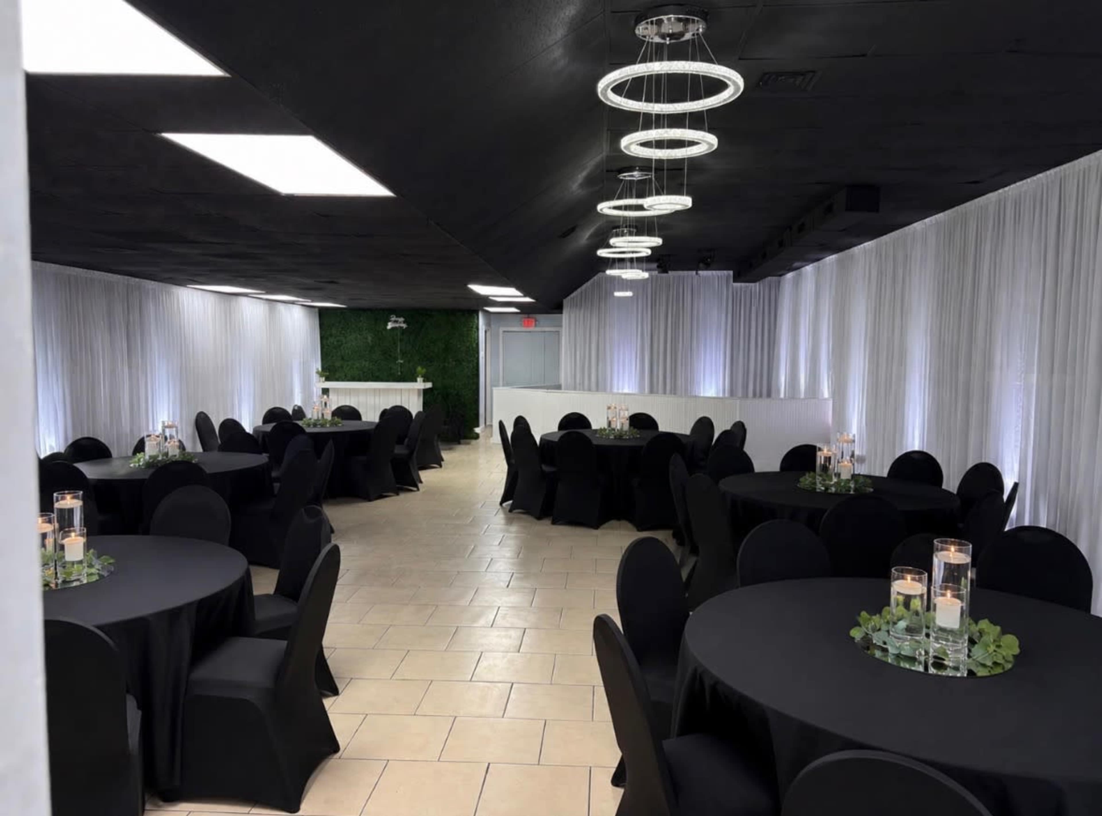 The image depicts a well-lit event space featuring multiple round tables covered with black tablecloths and surrounded by black chairs, with a green wall backdrop and subtle centerpieces of candles and greenery.