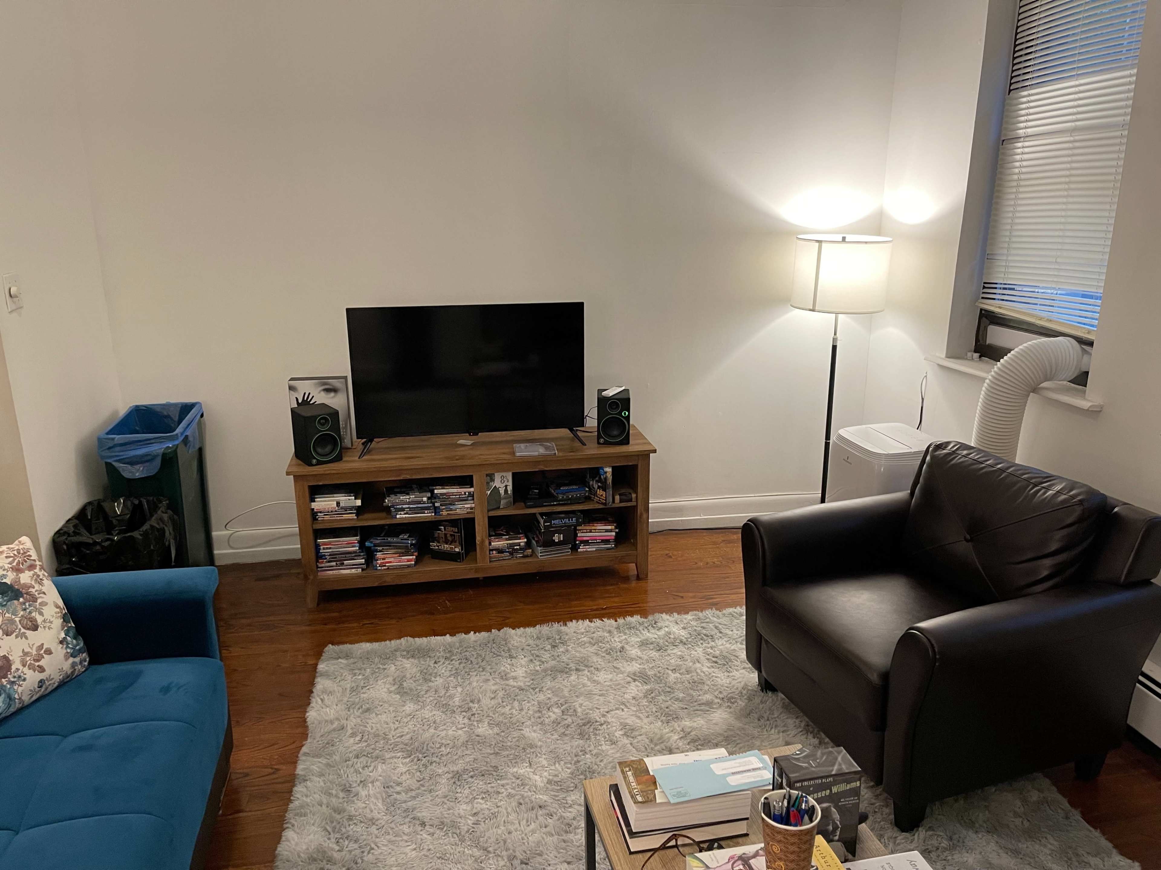 The image shows a living room with a blue couch, a black armchair, a wooden TV stand with speakers, a television, and a floor lamp, along with a trash can and a portable air conditioner in the corner.