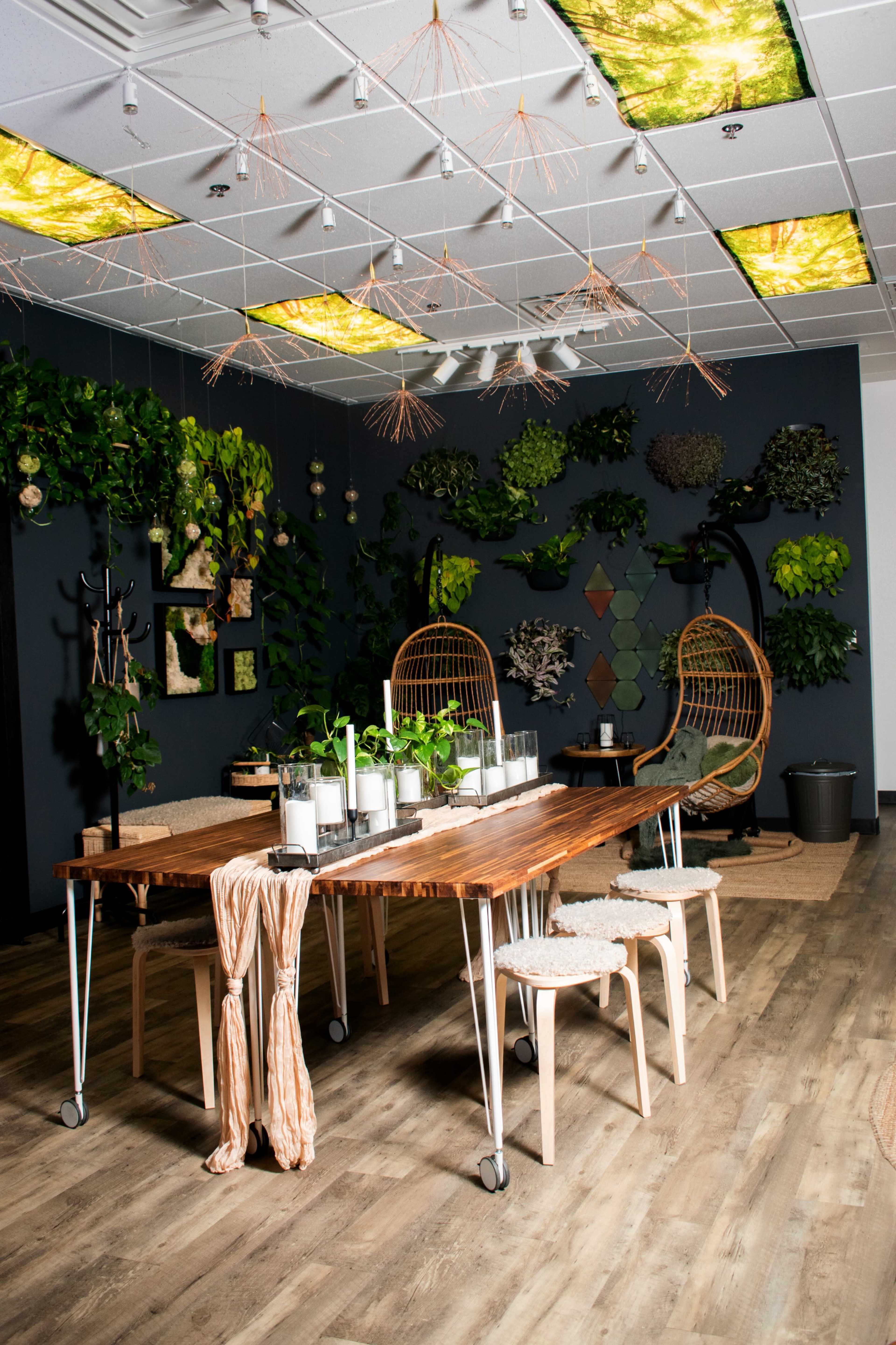 A modern dining area features a long wooden table with a central plant arrangement, surrounded by hanging chairs and walls adorned with greenery and decorative panels.