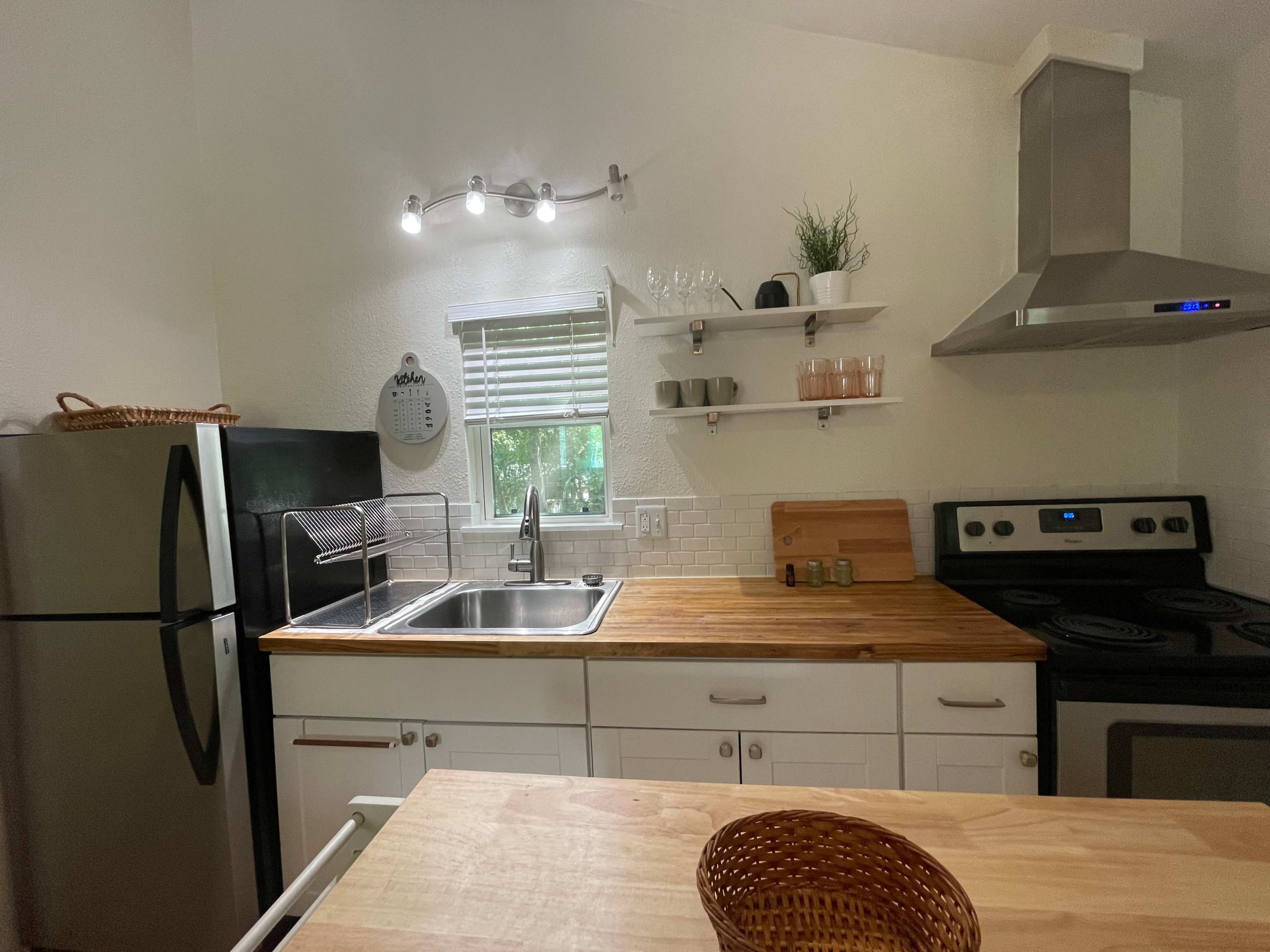 A compact kitchen features stainless steel appliances, a wooden countertop, and shelves with glassware and plants.