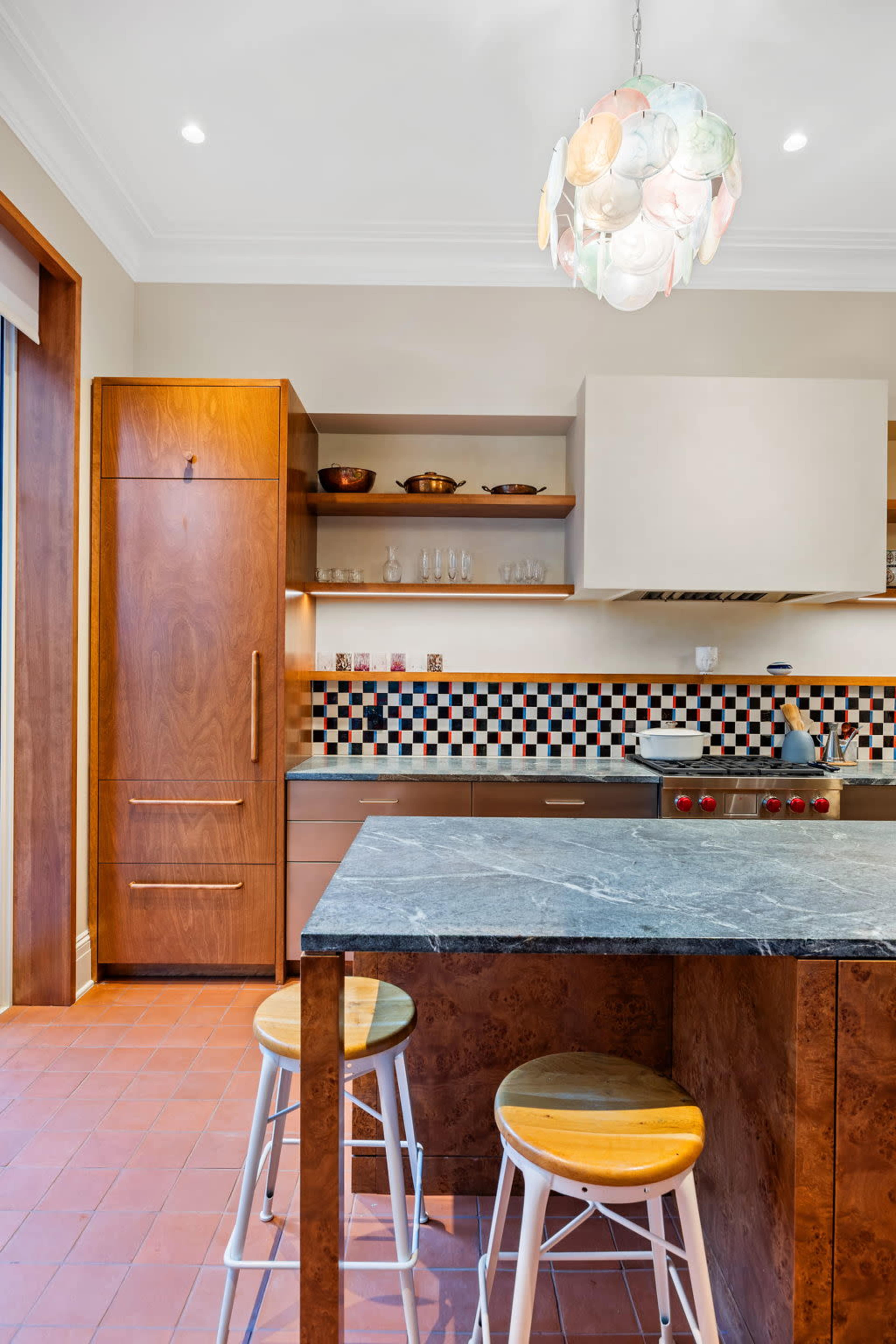 The image shows a modern kitchen with wooden cabinetry, a marble countertop, and a colorful backsplash featuring black, white, and orange tiles, complemented by a unique chandelier.