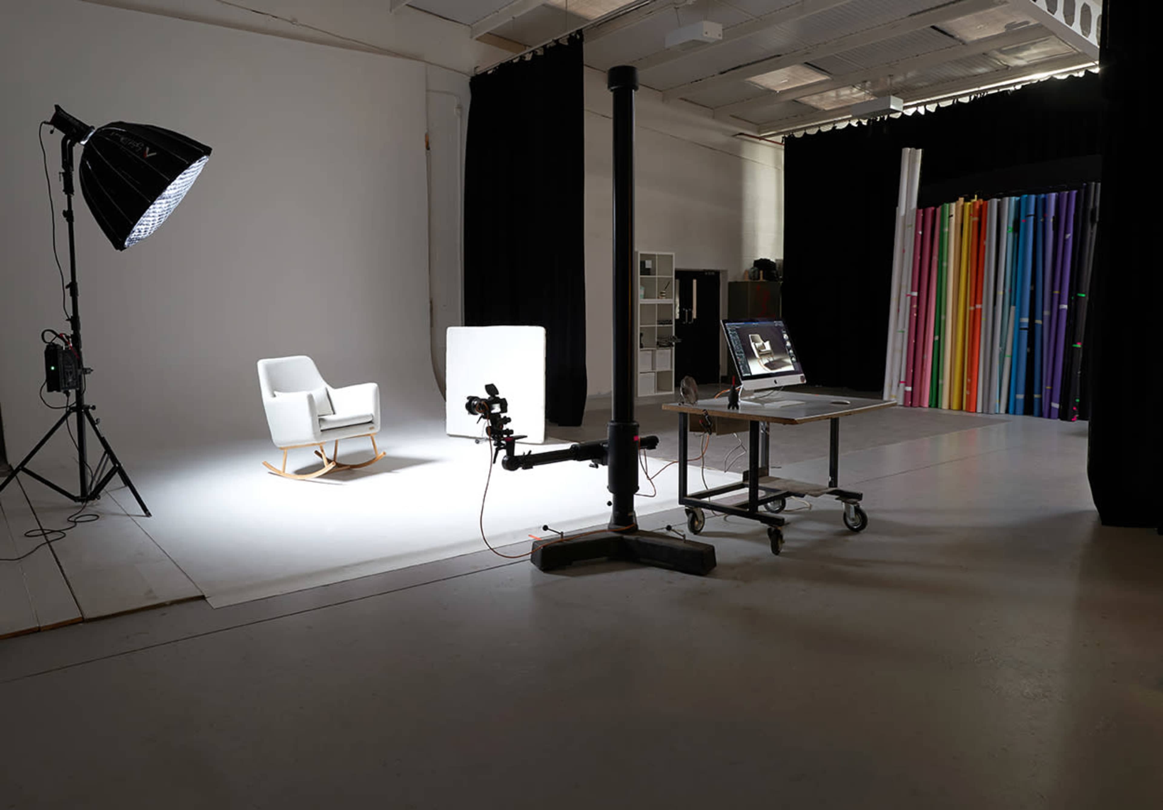 A photography studio features a white backdrop, a rocking chair, a large light source, and a mobile workstation with a computer.