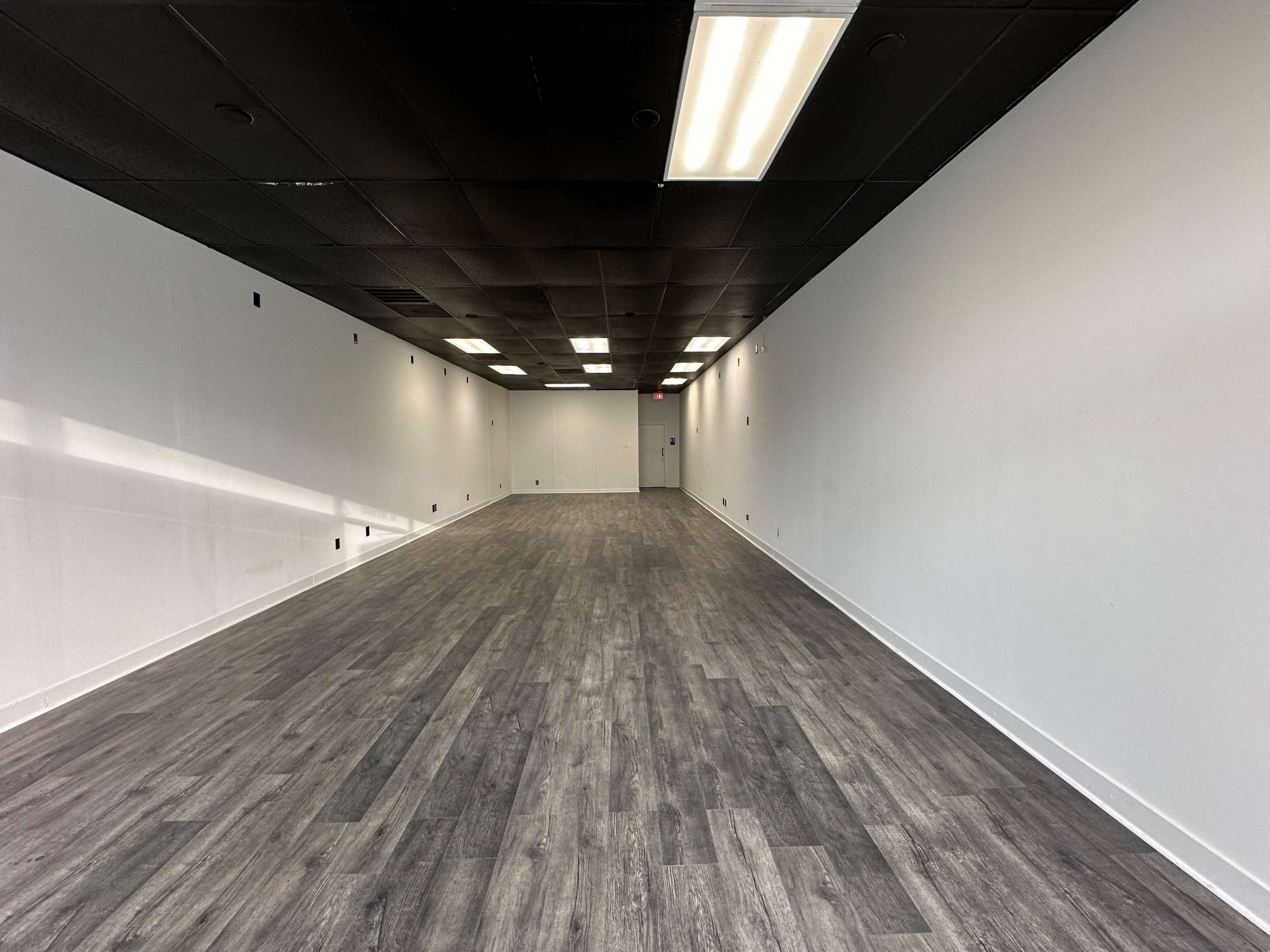 A blank, empty room with a polished wood-like floor and white walls, illuminated by fluorescent lighting.