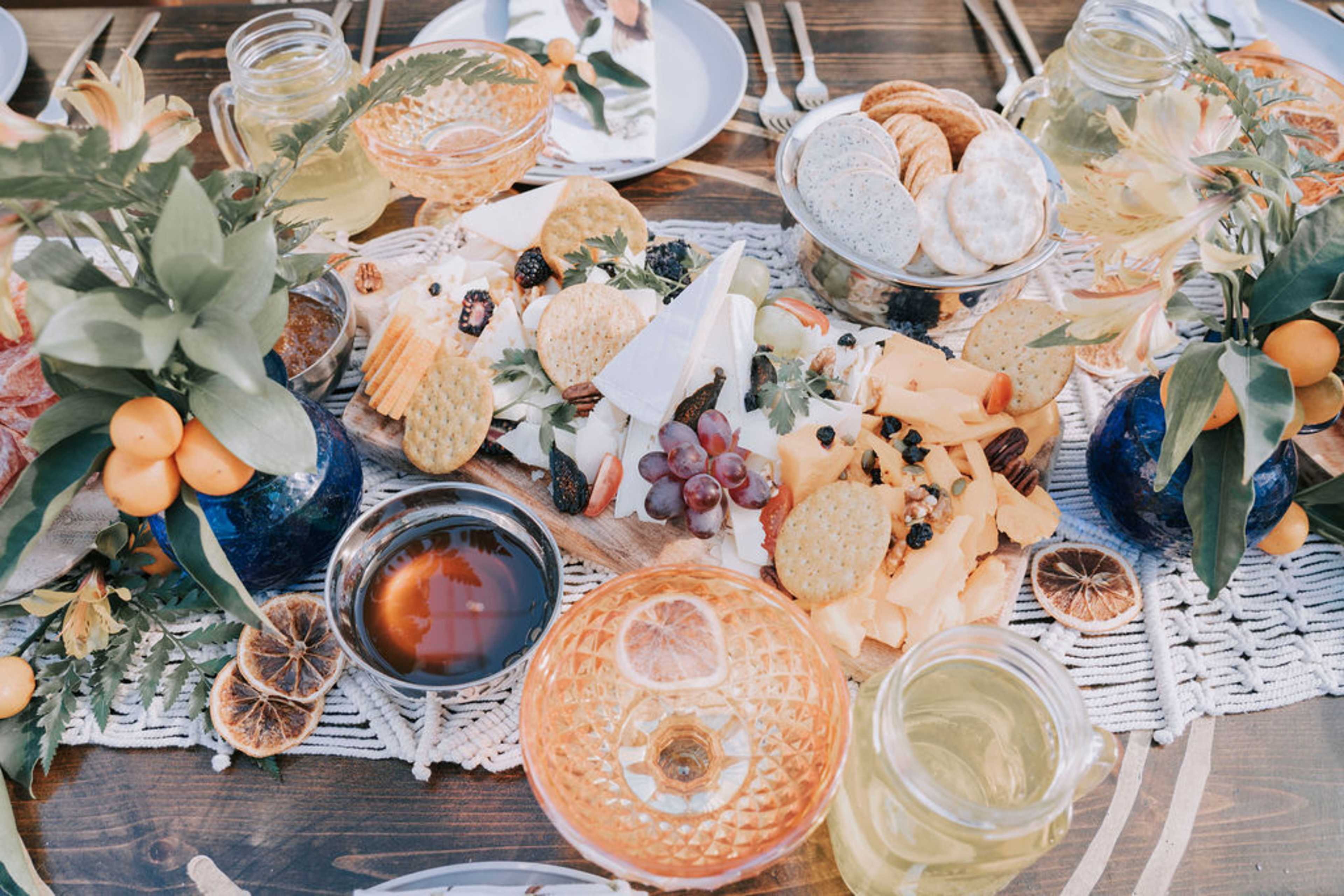 The image features a large wooden cheese board surrounded by various cheeses, crackers, fruits, and drinks on a beautifully arranged dining table.