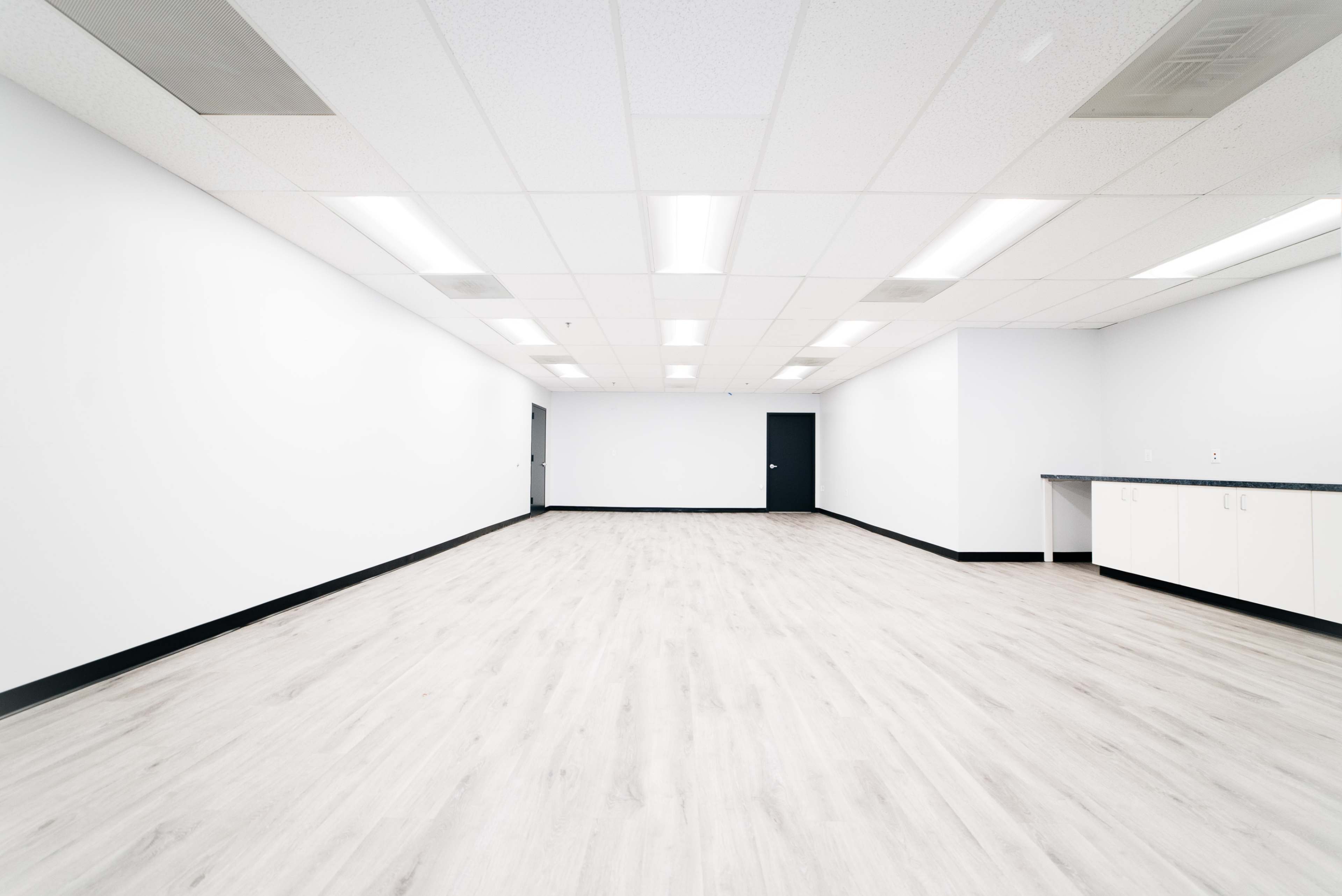 The image shows a brightly lit, empty room with light-colored walls, a wooden floor, and a dark door and counter along one side.