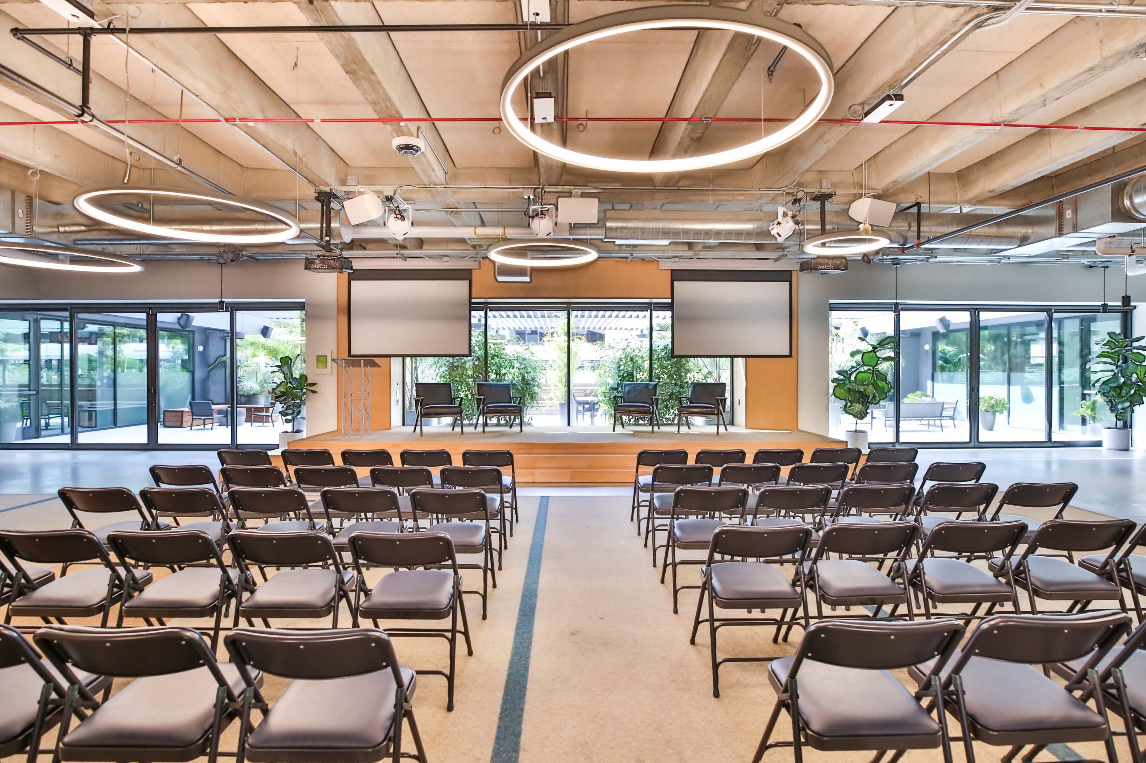 A modern conference room features a stage with chairs, two large screens, and circular light fixtures above, surrounded by glass walls and plants.