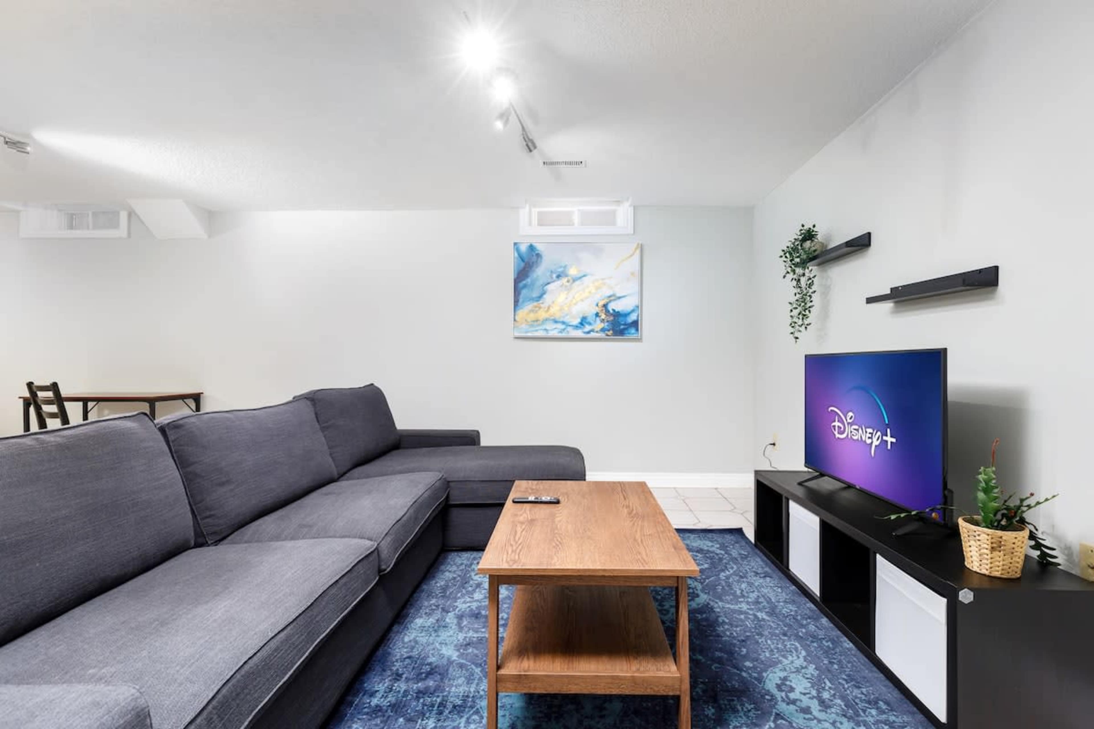 A modern living room features a gray sectional sofa, a wooden coffee table, a television displaying the Disney+ logo, and a painting on the wall.