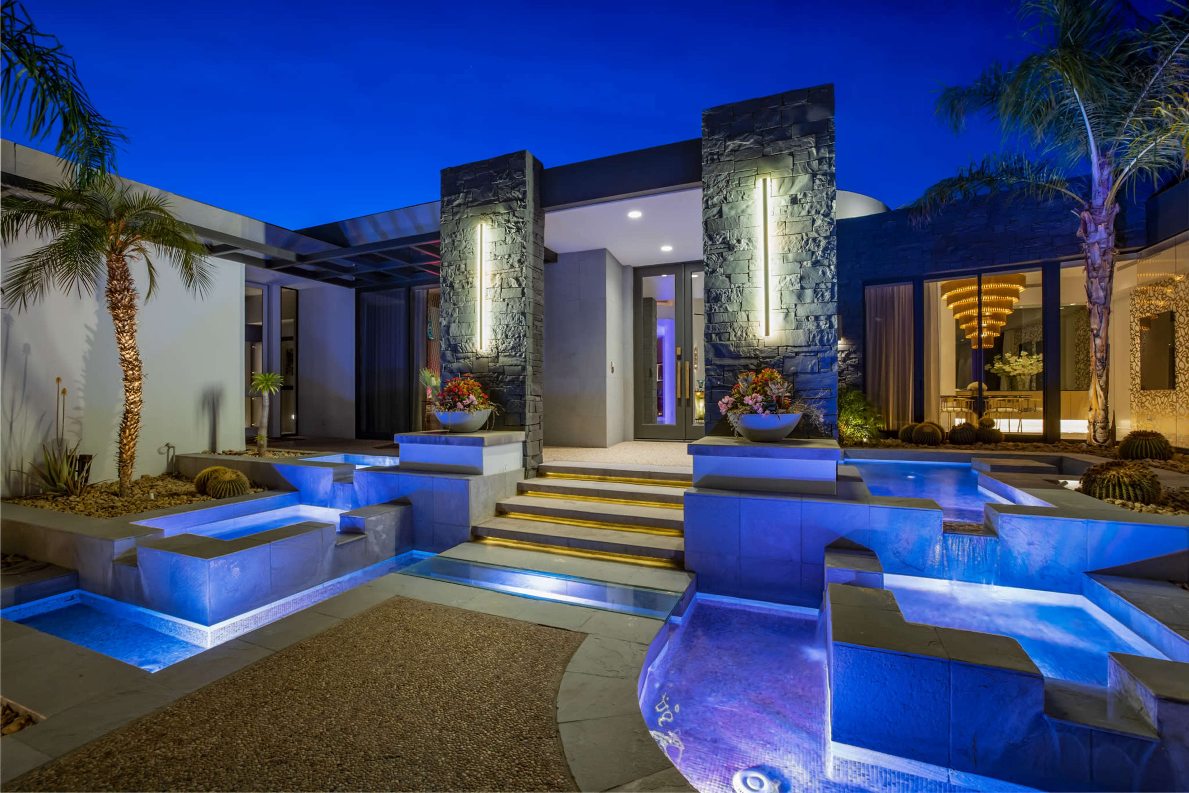 The image shows a modern home entrance with illuminated steps, landscaped planters, and water features under a twilight sky.