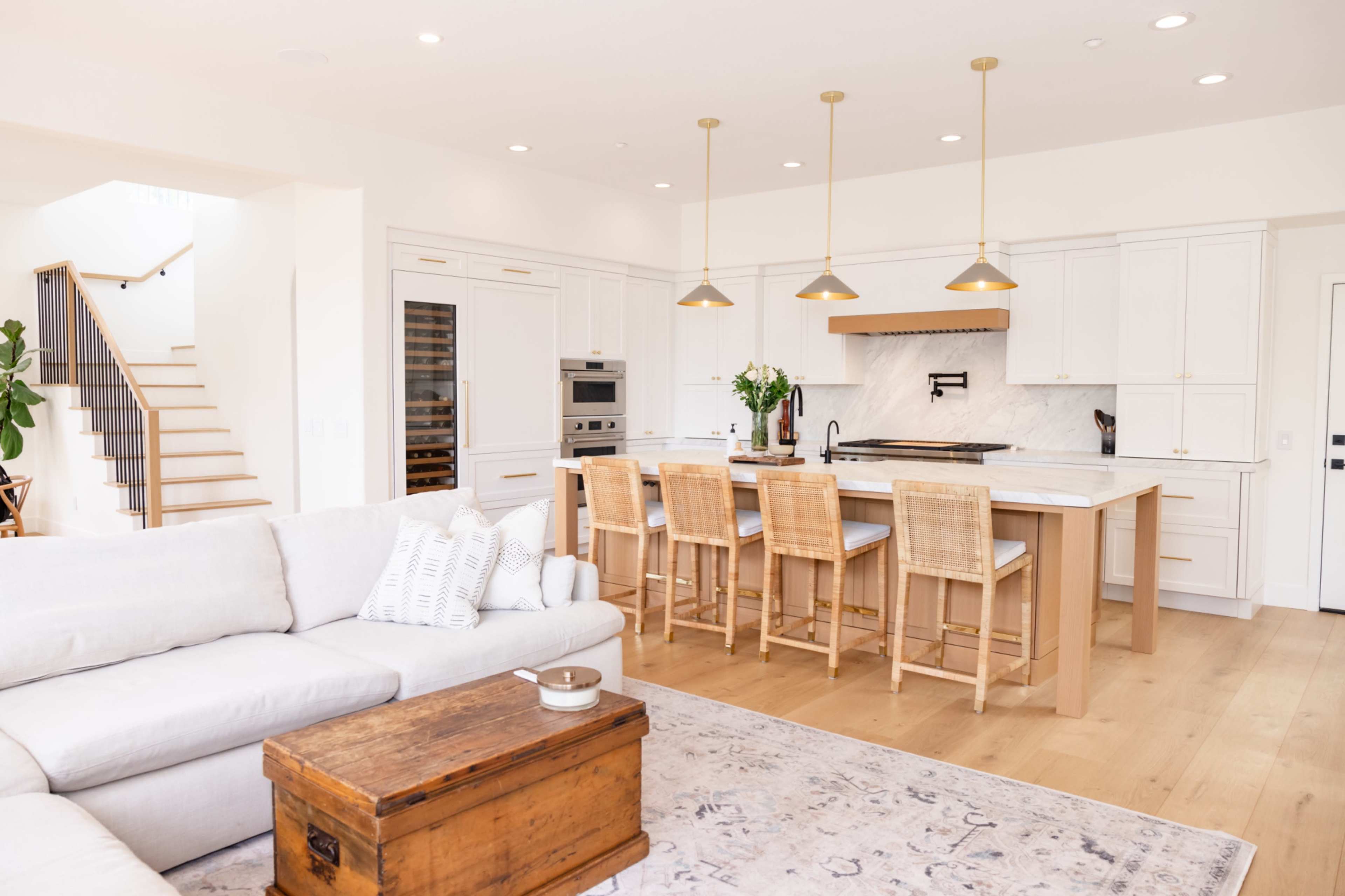 The image shows a modern kitchen with a large island, three pendant lights overhead, and bar stools, adjacent to a living area featuring a light-colored sofa and a wooden coffee table.