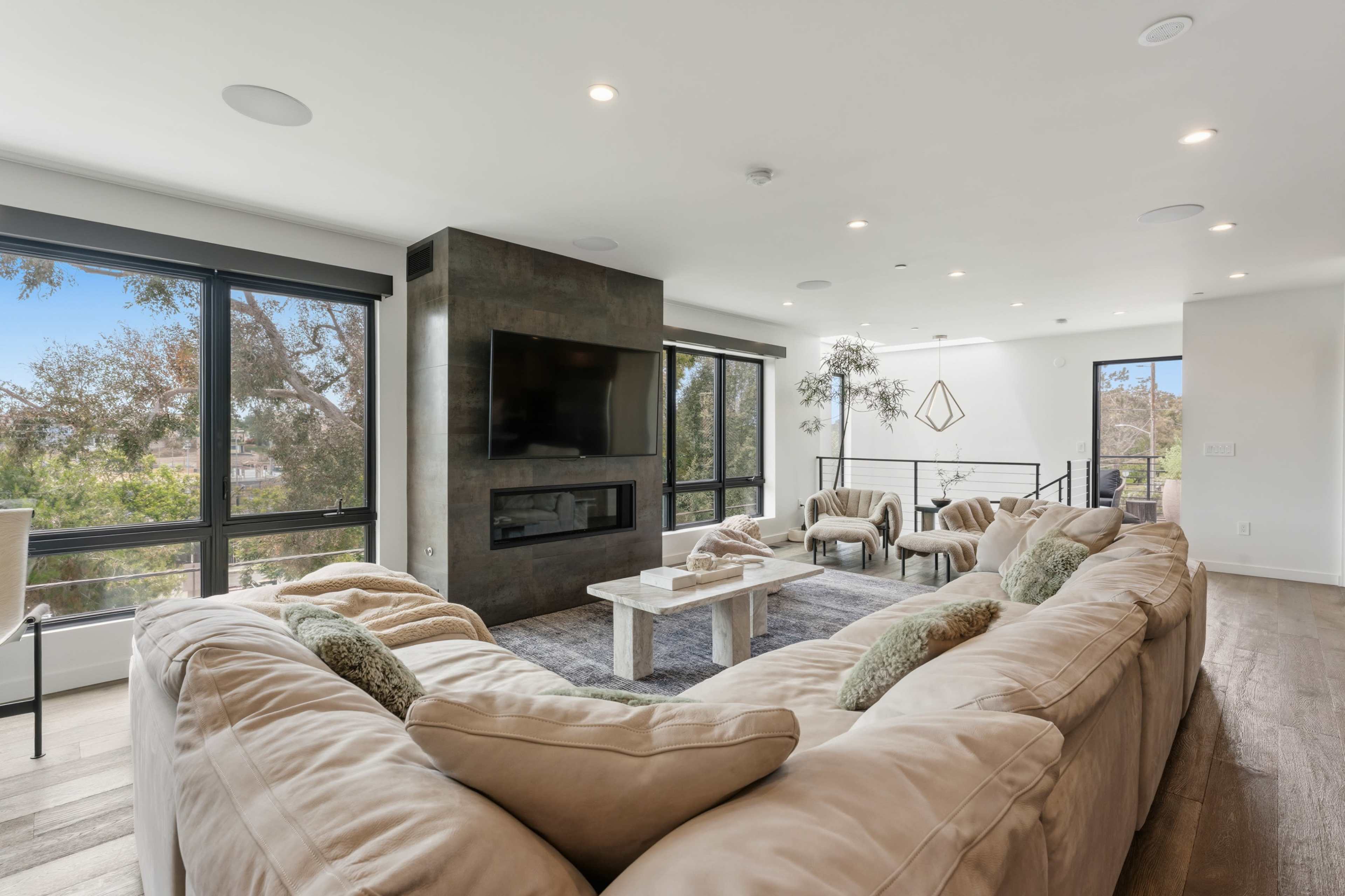 A spacious living room features a large sectional sofa, a stone fireplace, and expansive windows offering views of outdoor greenery.