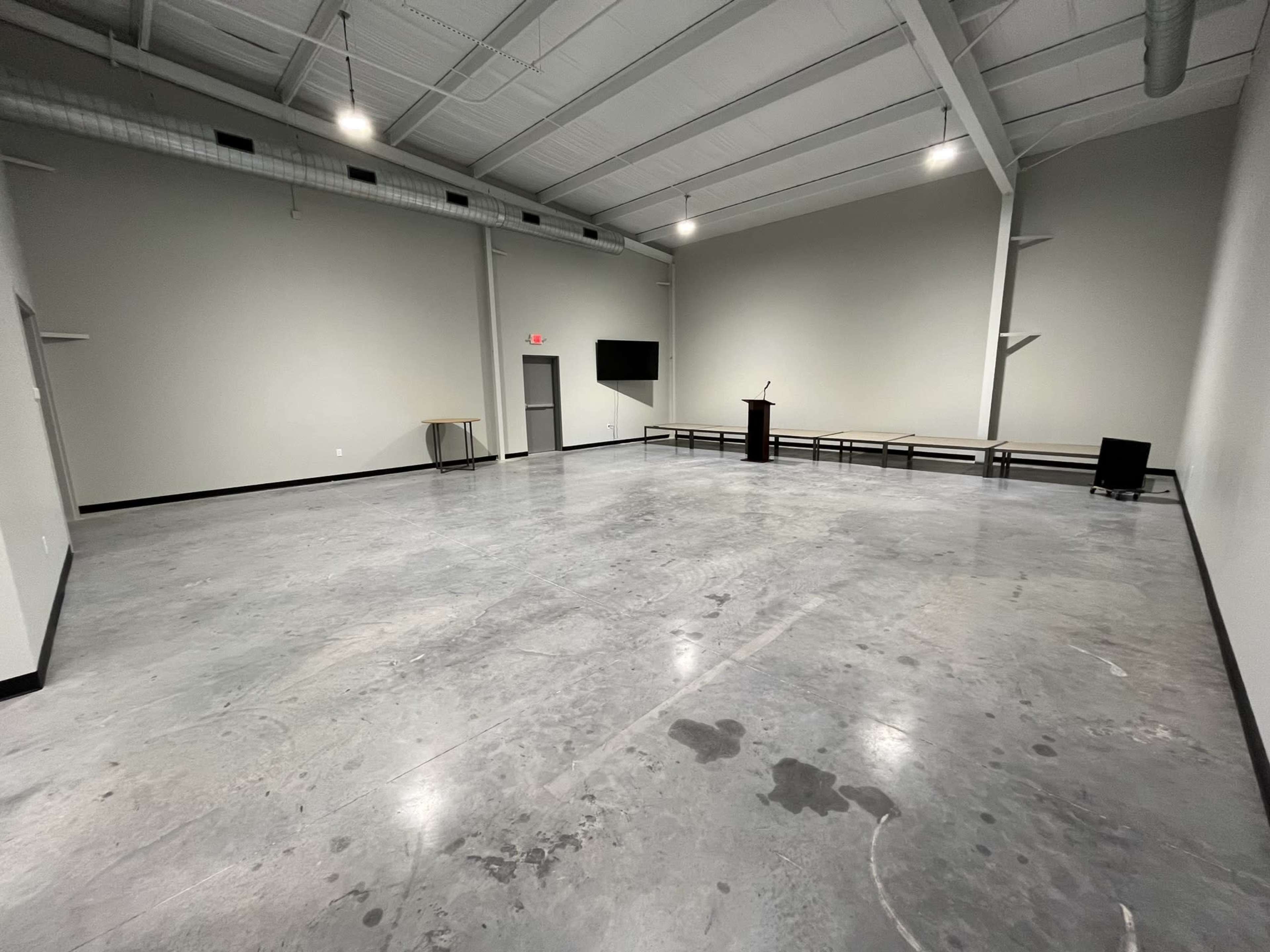 A spacious, empty interior room with a polished concrete floor, a podium at one end, and minimal furnishings against the walls.