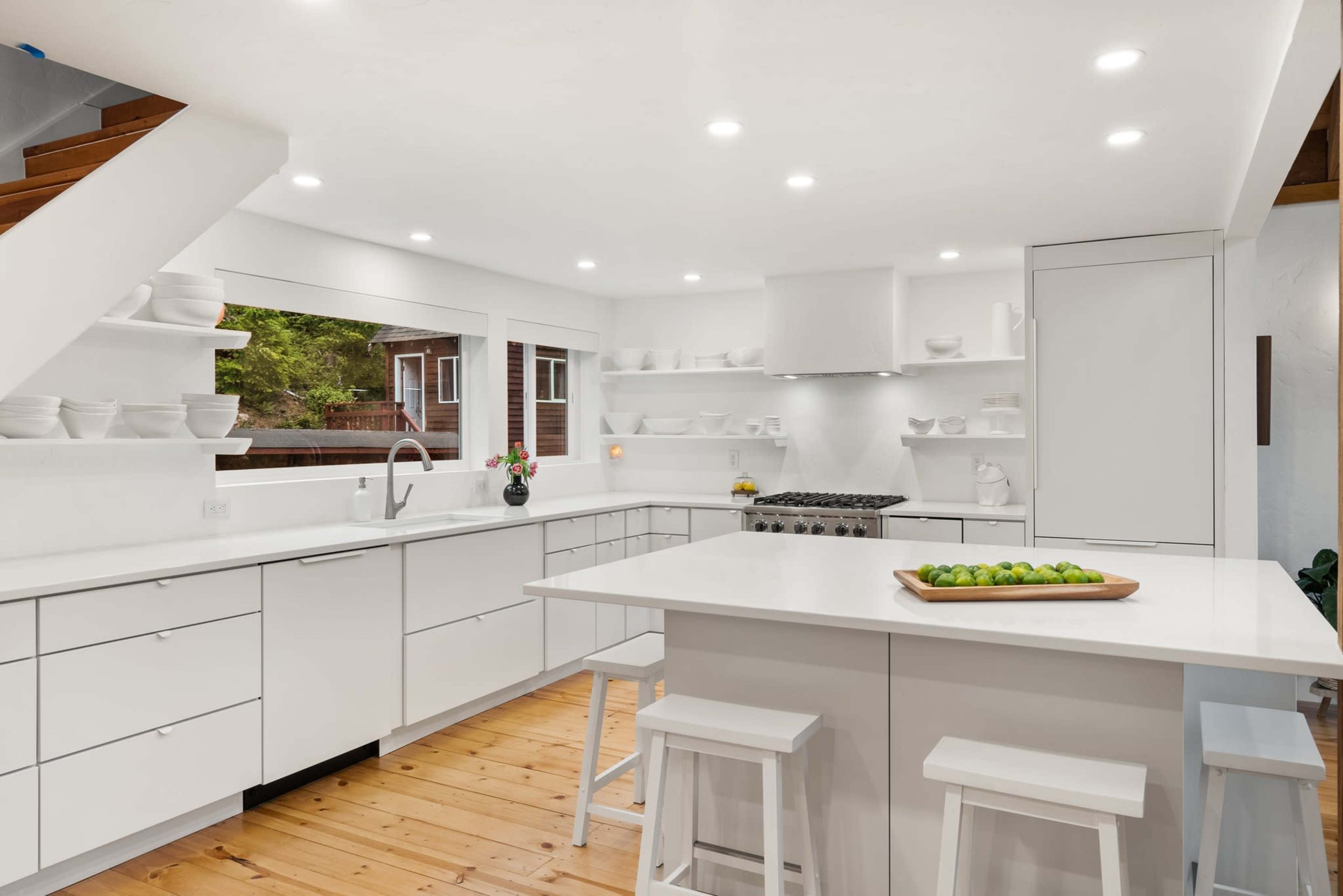 The kitchen features white cabinetry, a central island with a wood tray of limes, and large windows that provide natural light.