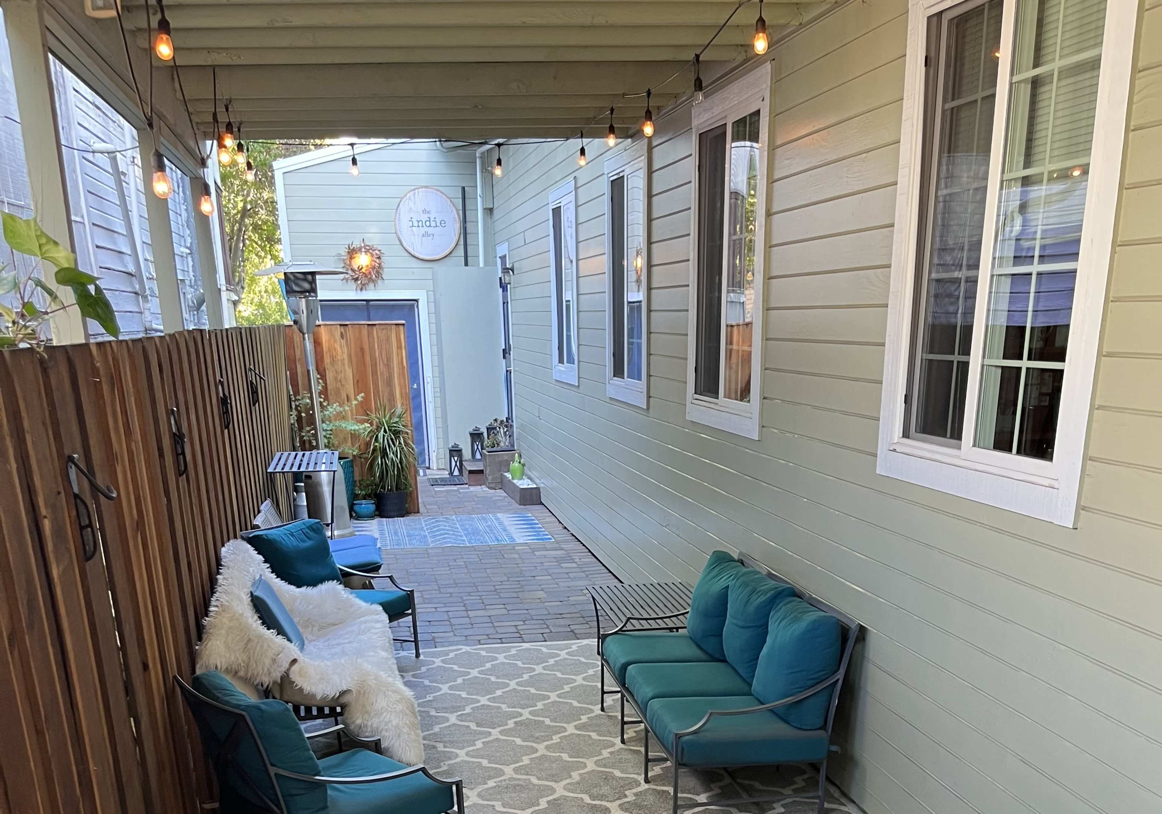 A narrow outdoor seating area lined with green walls, featuring two teal chairs, a rug, and string lights overhead.
