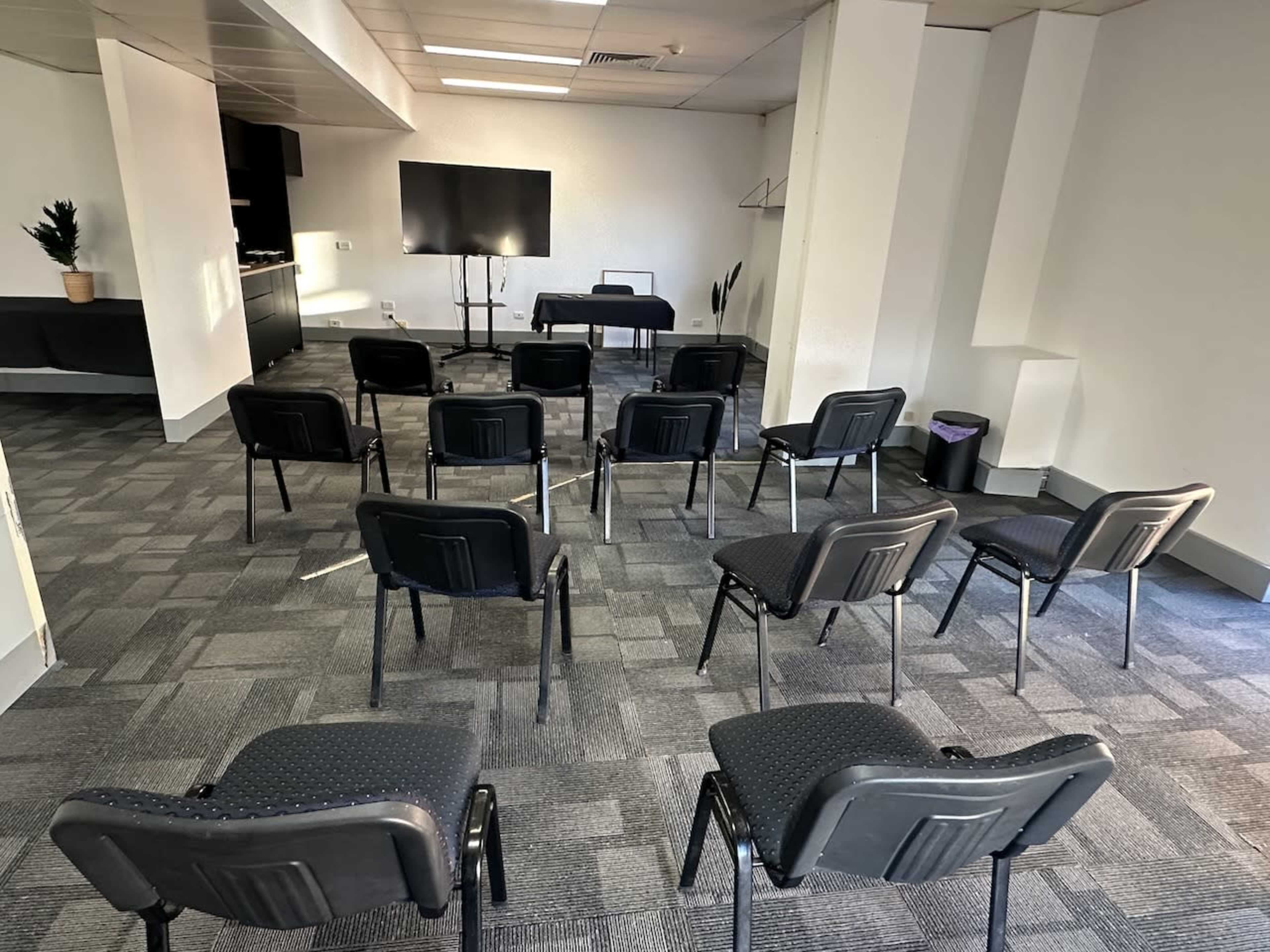 A conference room is set up with several black chairs arranged in rows facing a table and a large screen.