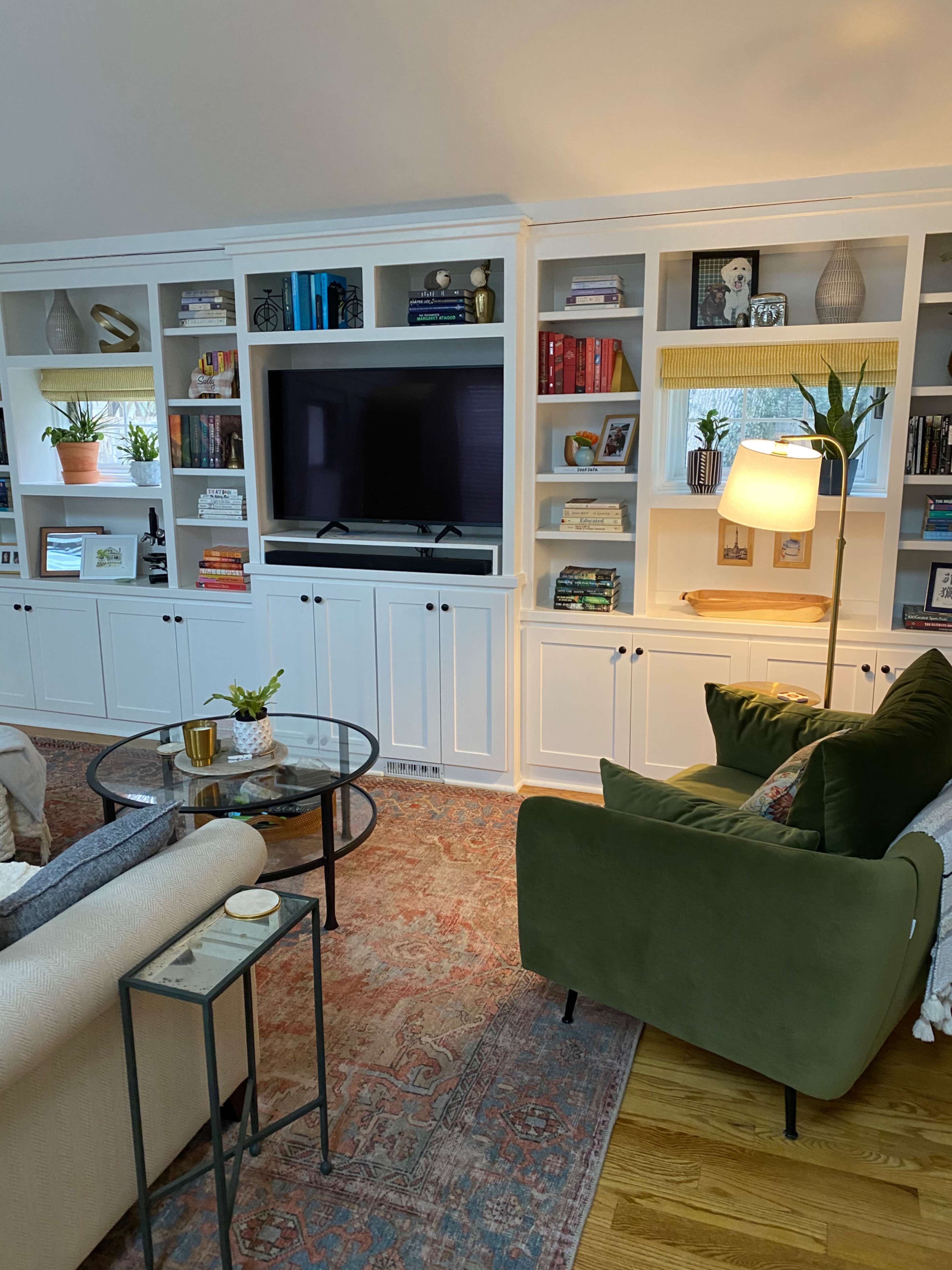 The image shows a cozy living room with built-in shelves featuring books and decor, a television in the center, and a green armchair next to a small table.