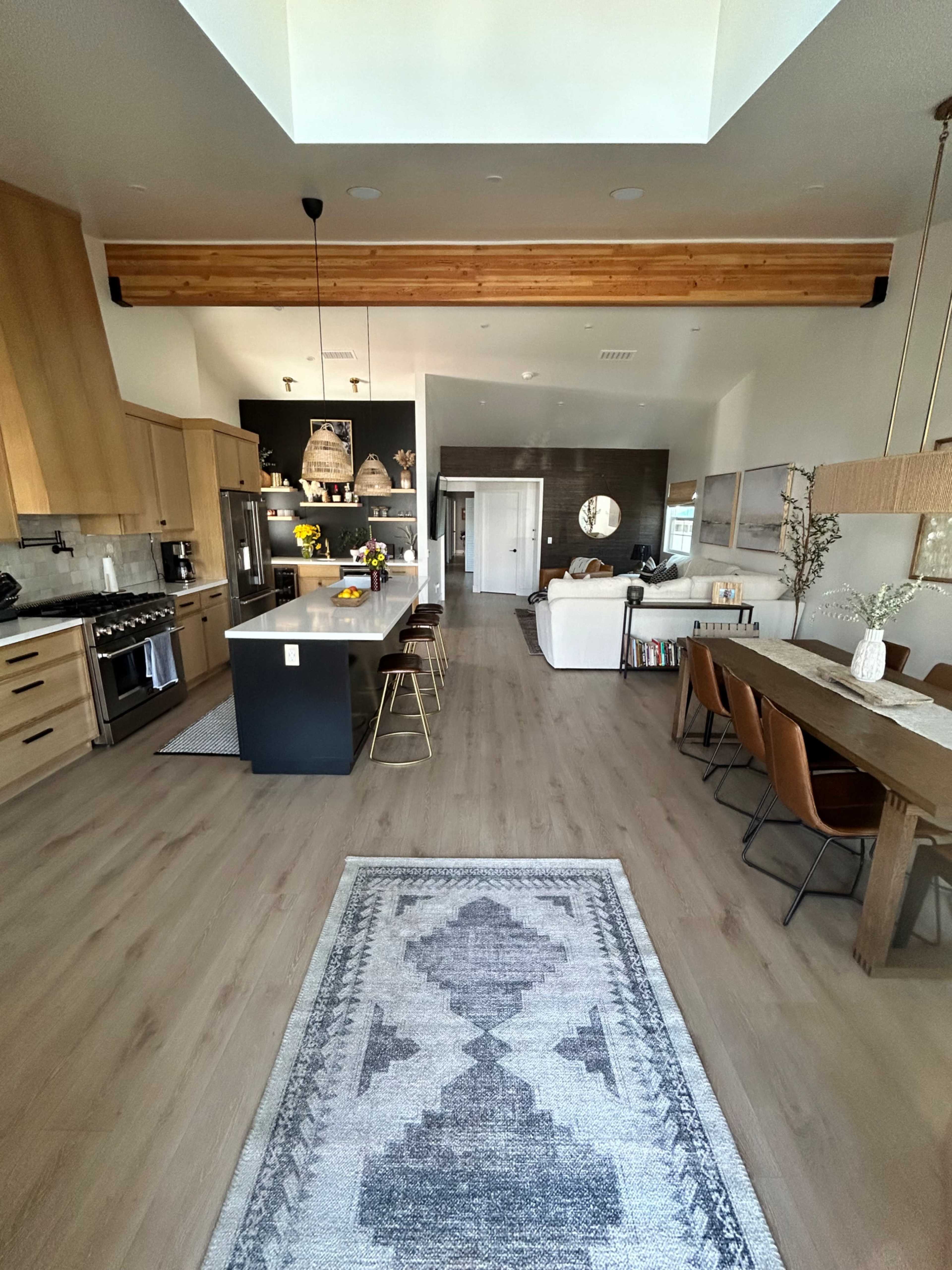 A spacious modern kitchen and dining area features wood accents, a central island with seating, and a view of a living room with a large window and comfortable furniture.
