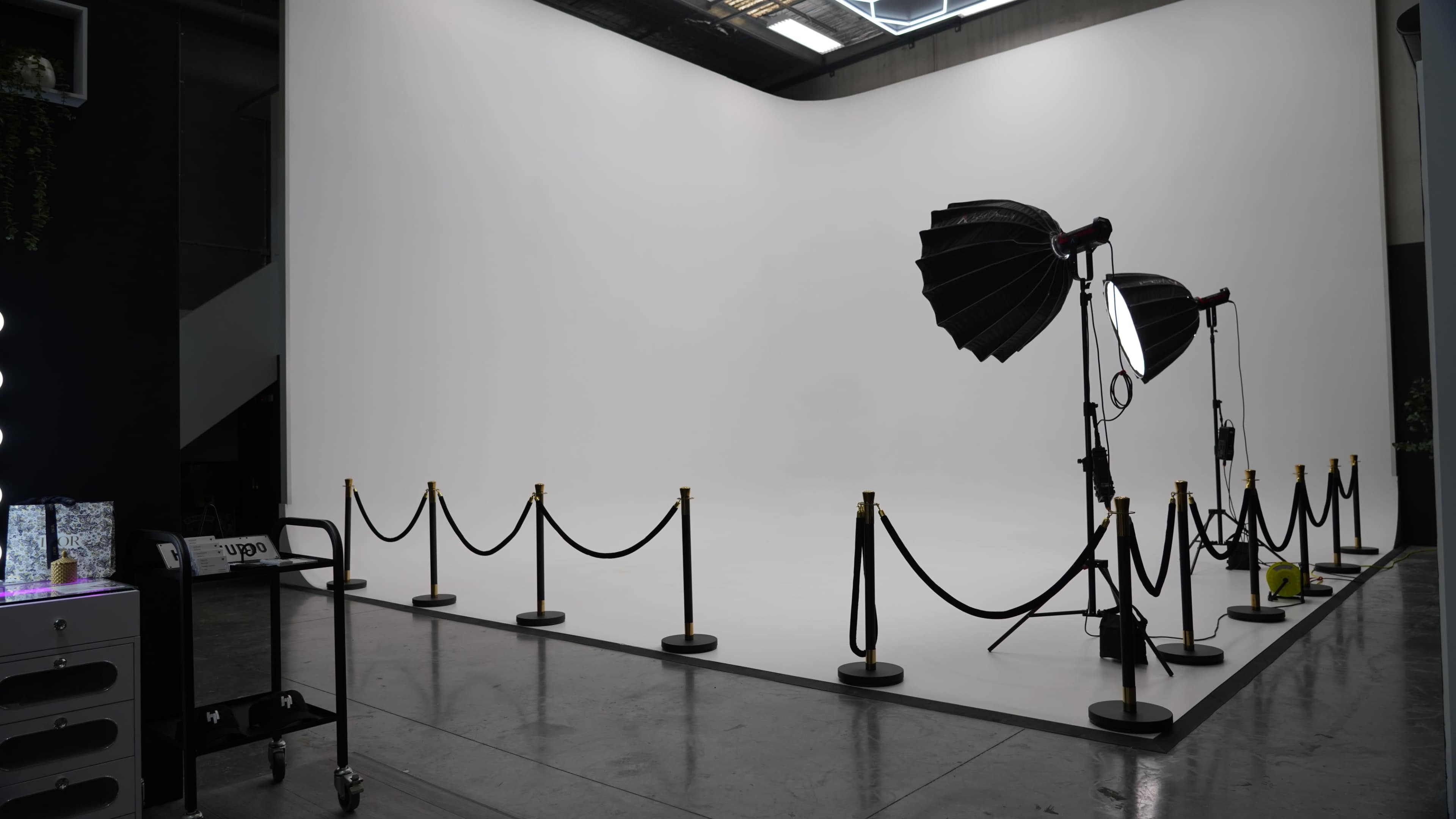 The image shows a minimalist photography studio with a blank white backdrop, lighting equipment, and a rope barrier.