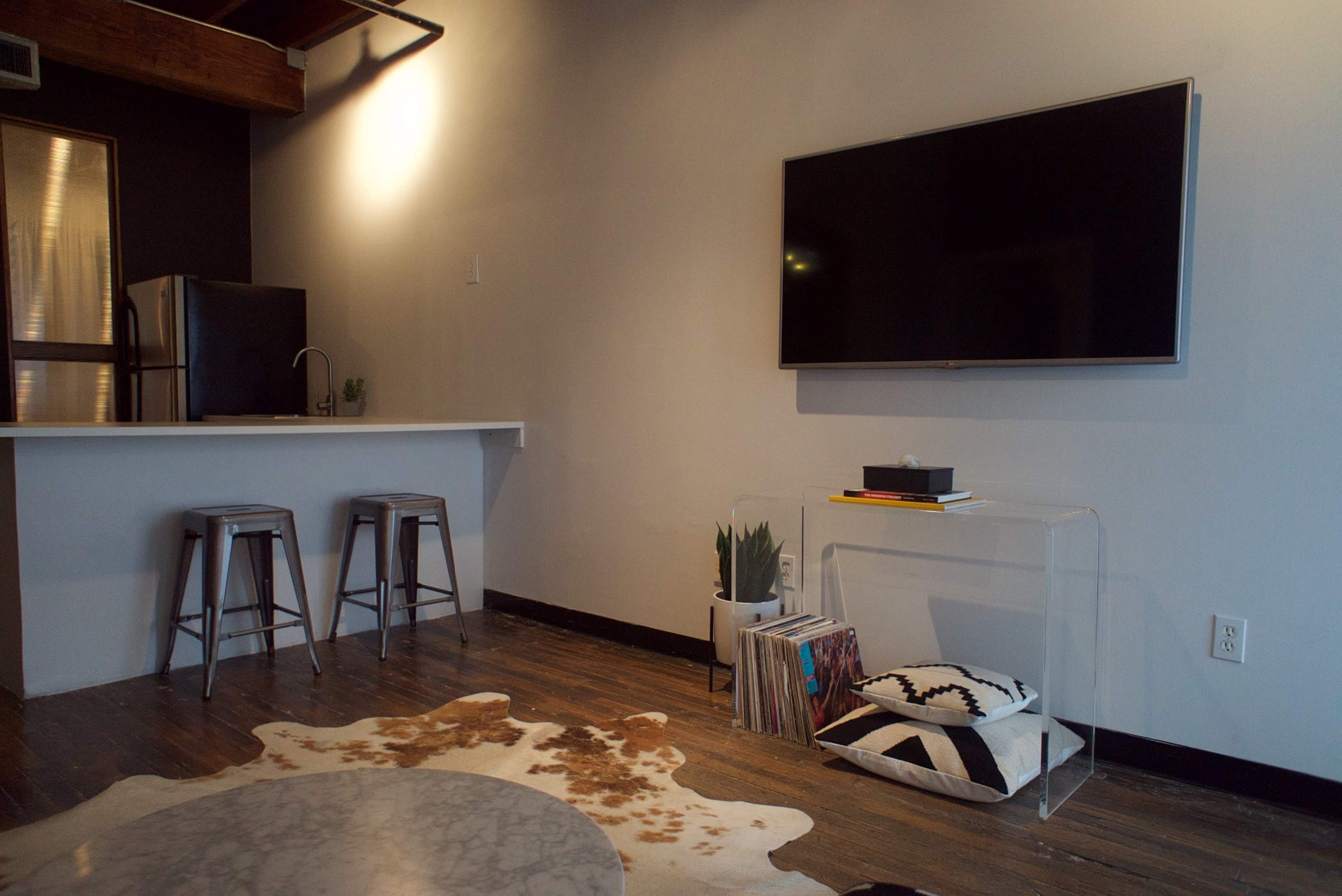 The image shows a modern living space featuring a television mounted on the wall, a clear coffee table with books, a bar counter with two stools, and a cowhide rug on a wooden floor.
