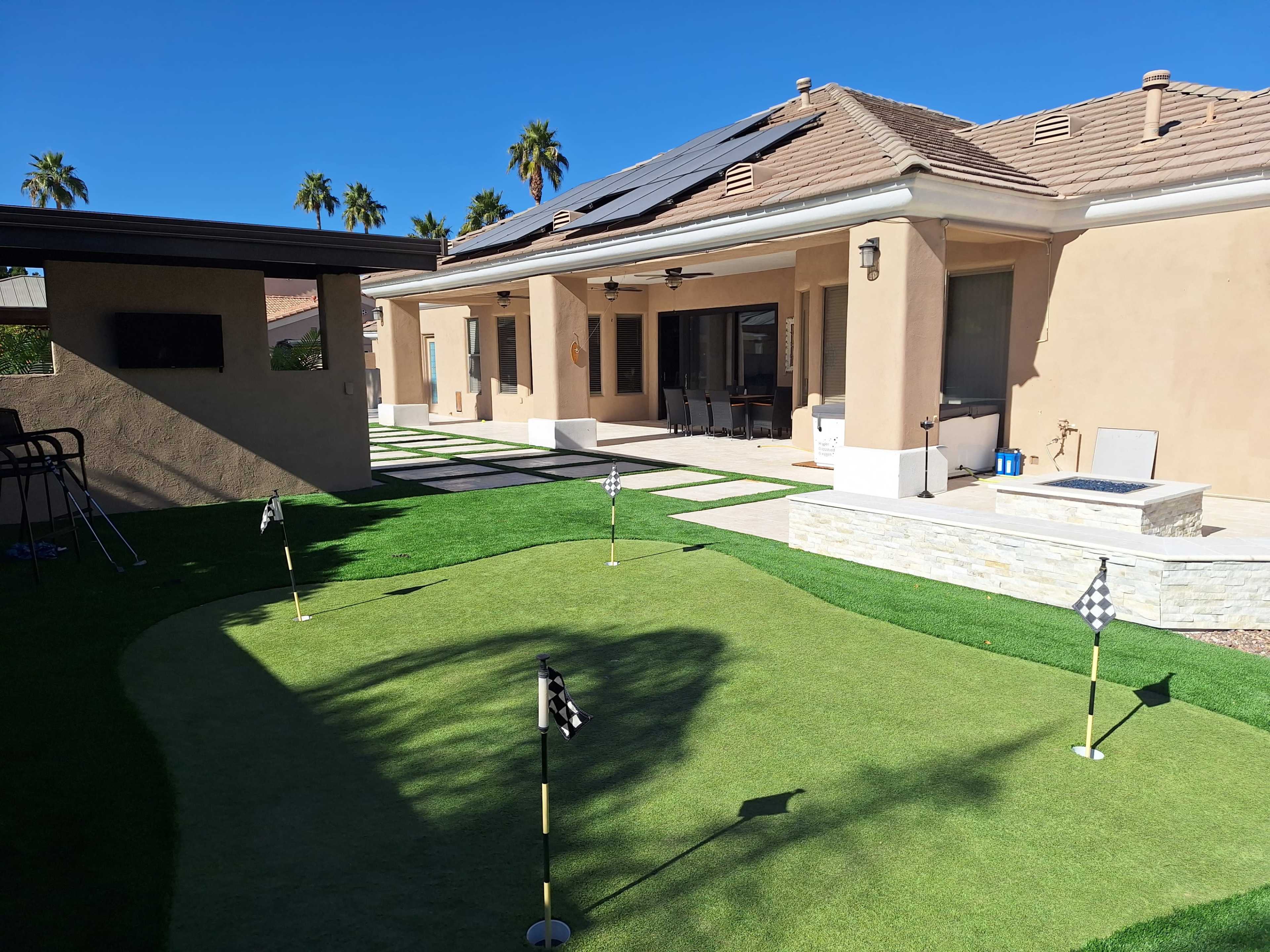 A backyard features a putting green with flags, adjacent to a patio and a house with solar panels on the roof.