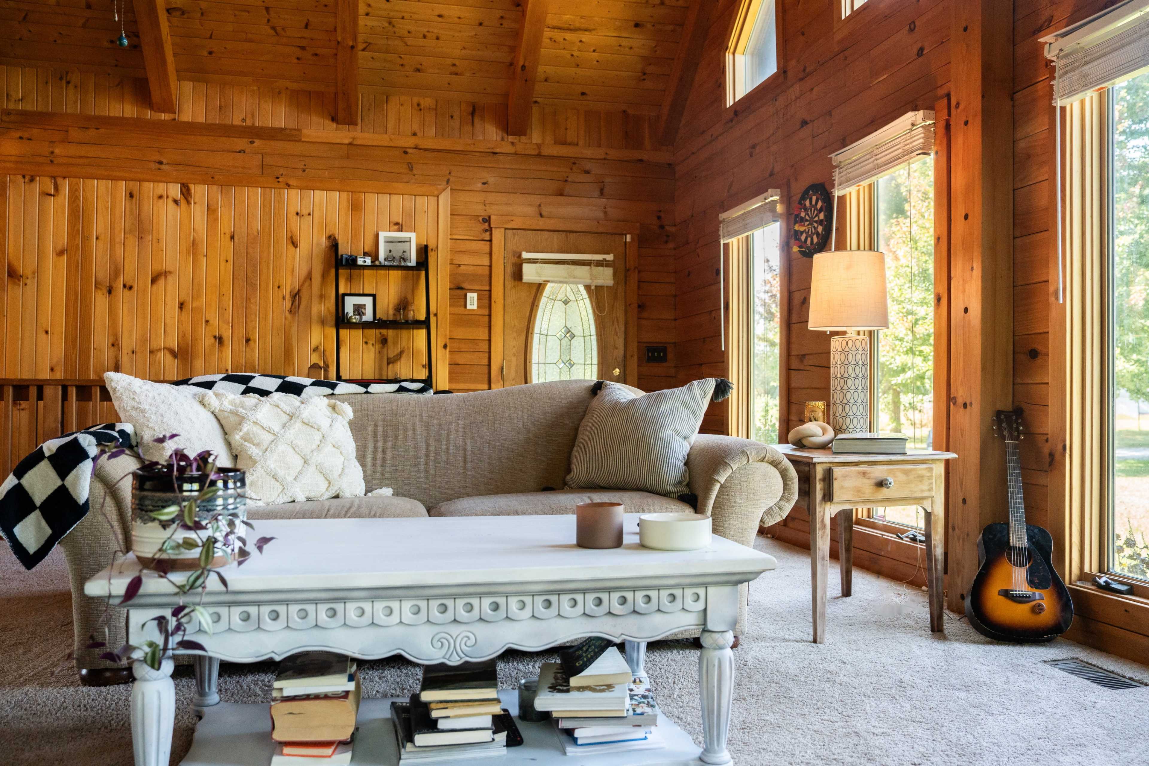 A cozy living room features a light-colored sofa, a white coffee table stacked with books, and large windows that illuminate the wooden interior.