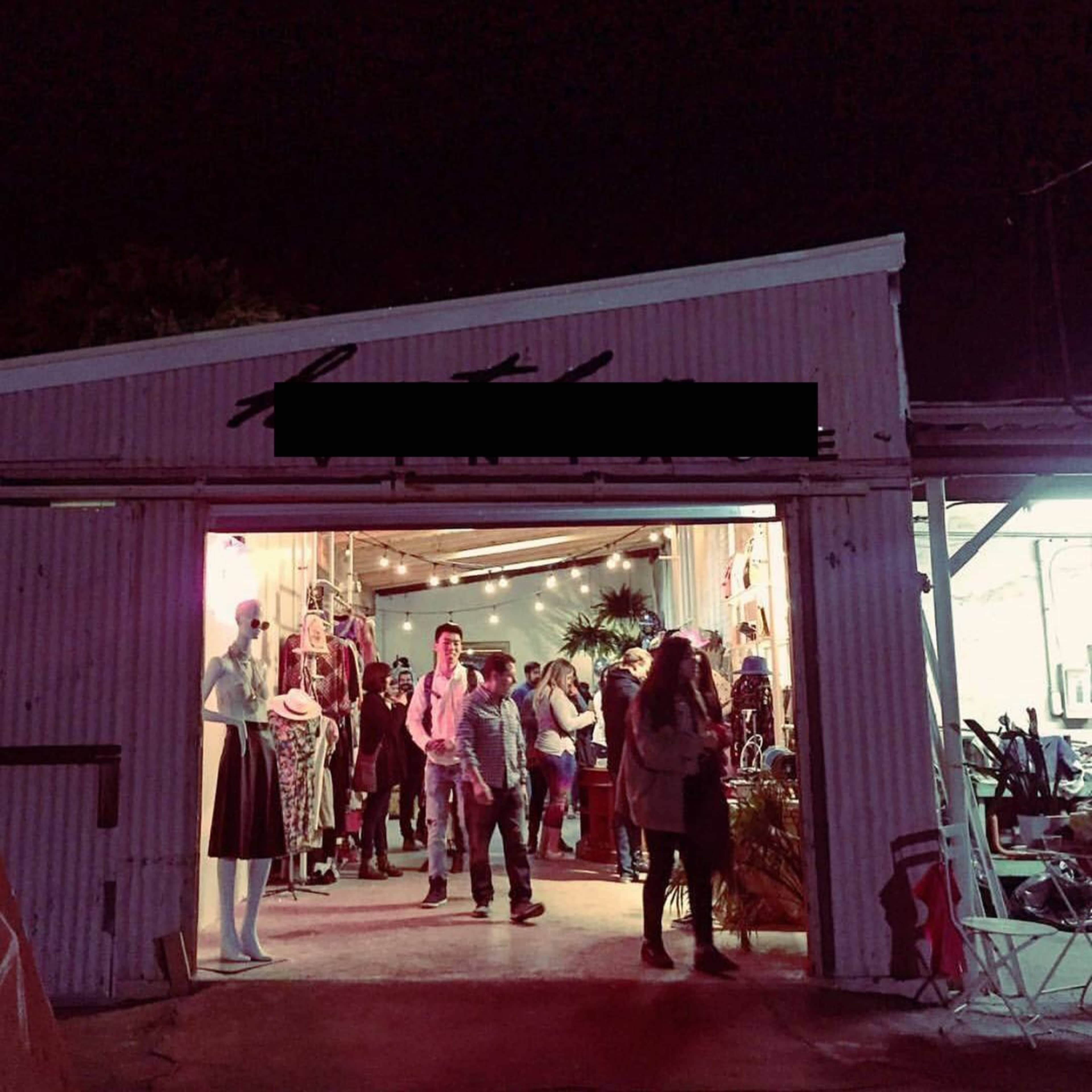 A group of people congregates outside a clothing store with a variety of garments displayed in the windows during the night.
