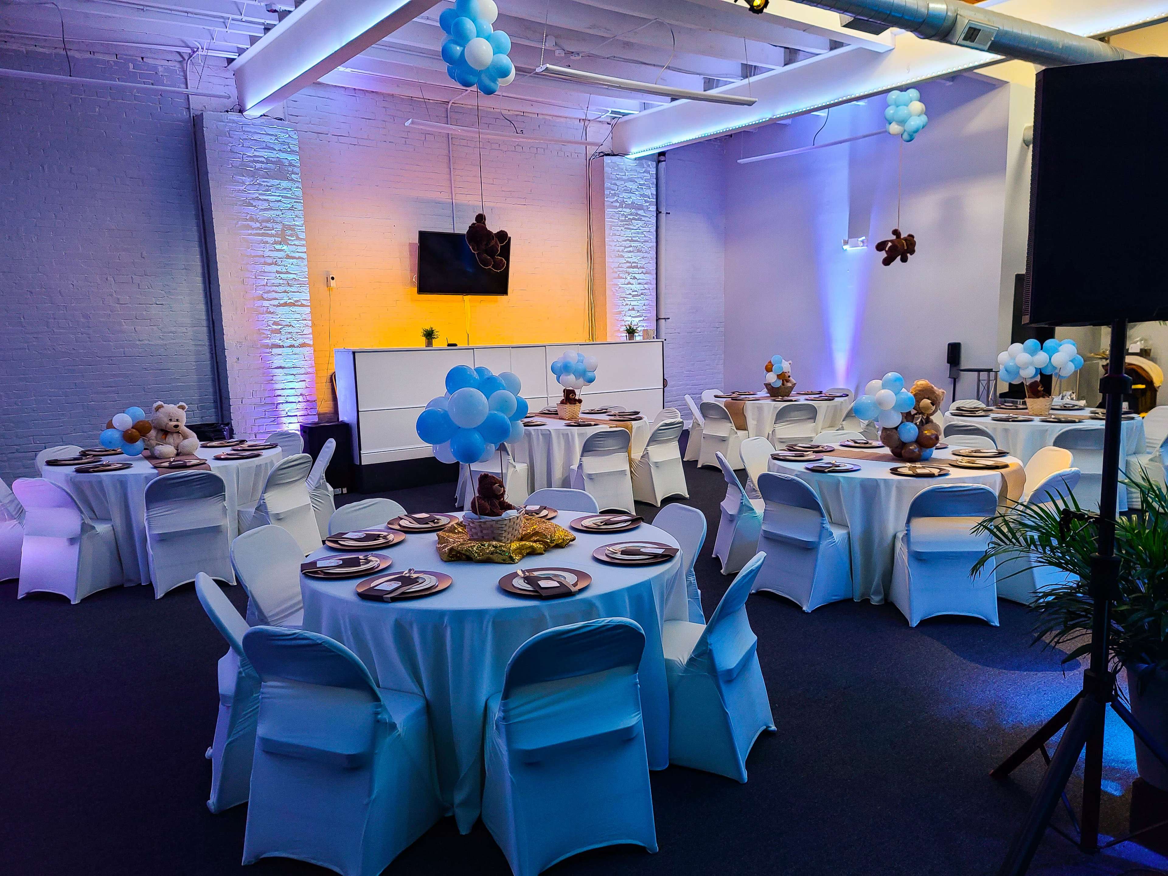 A spacious event room features round tables covered with white tablecloths, each adorned with plates and teddy bear centerpieces, set against a backdrop of blue balloons and colorful lighting.