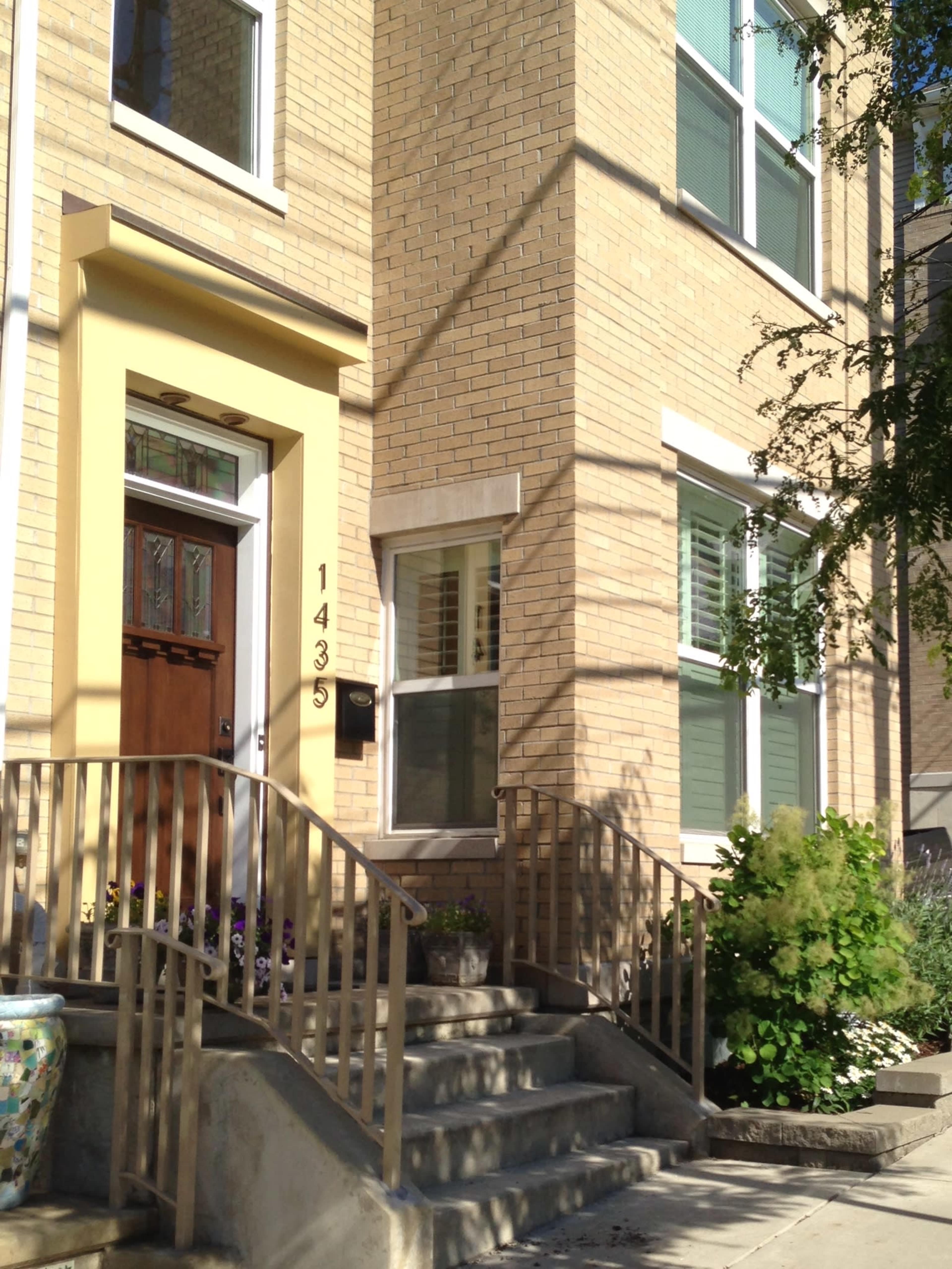 The entrance of a residential building with a yellow door, window shutters, and a set of concrete steps leading up to it.