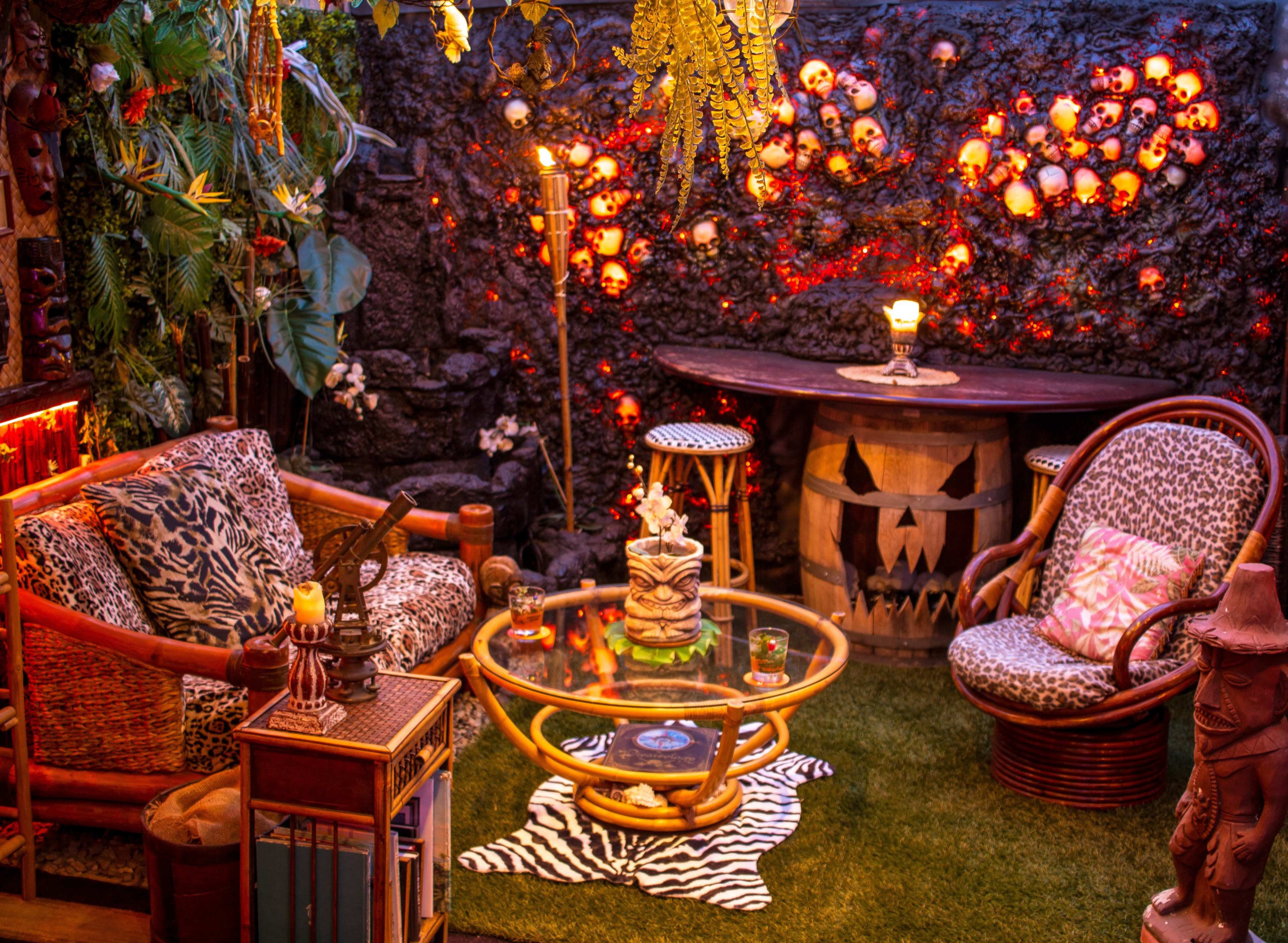 The scene features a cozy lounge area adorned with patterned furniture, a glass table, and decorative skulls on a textured wall, surrounded by tropical plants and warm lighting.
