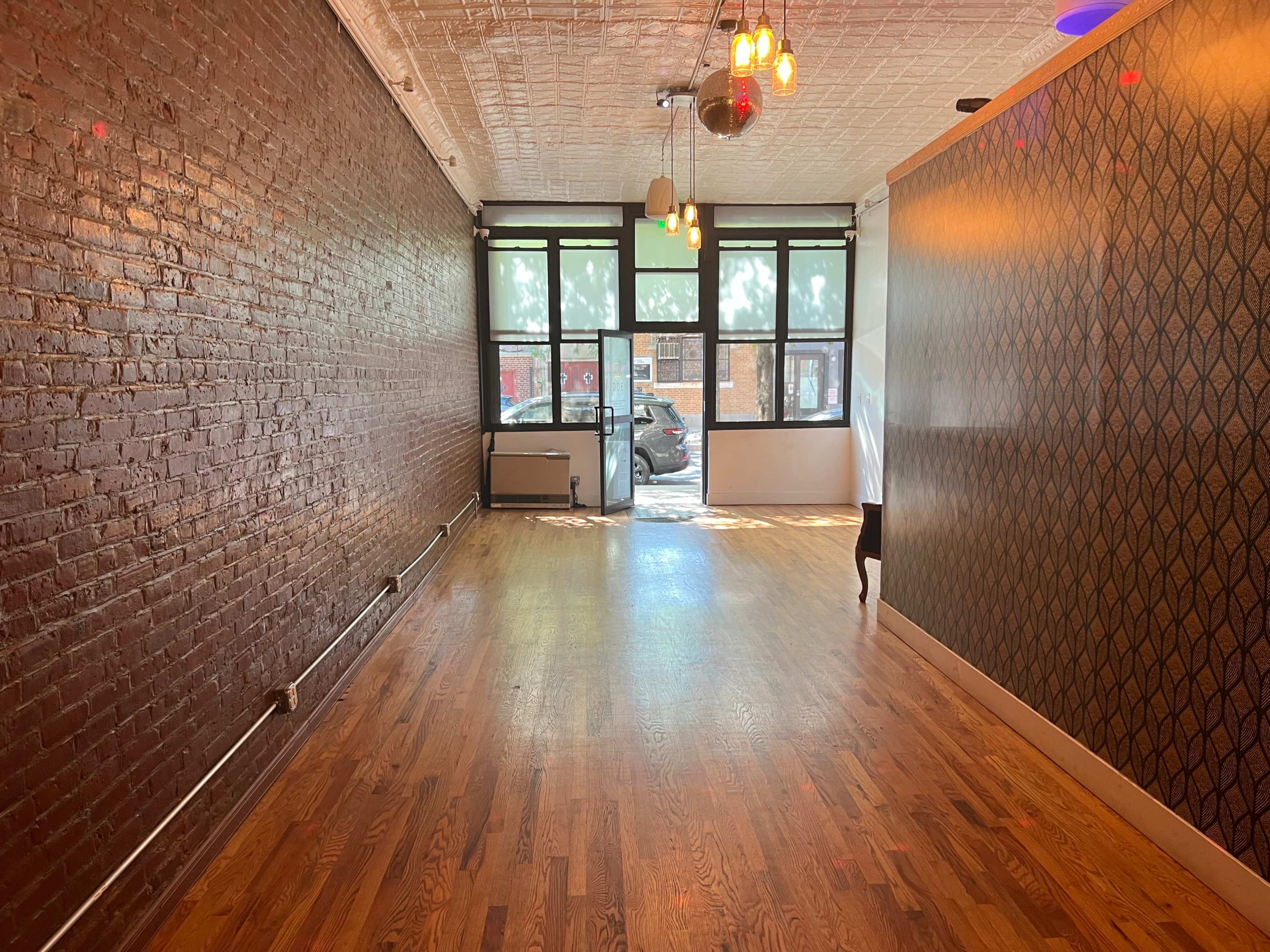 An empty room with exposed brick walls, hardwood flooring, and large windows allowing natural light to enter.