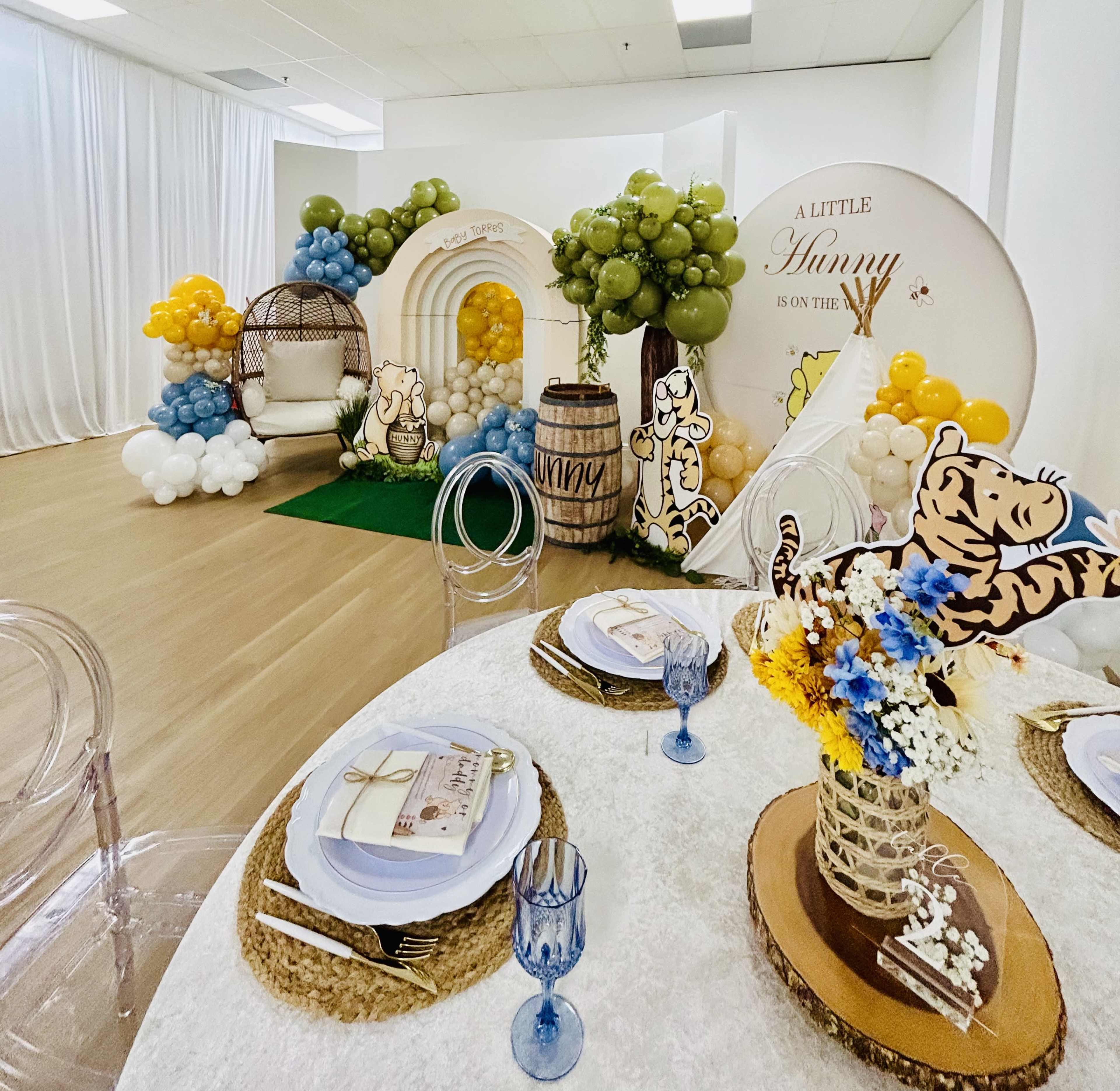 The image shows a decorated event space featuring a table set with plates and glasses, alongside balloon arrangements and themed decor in pastel colors.
