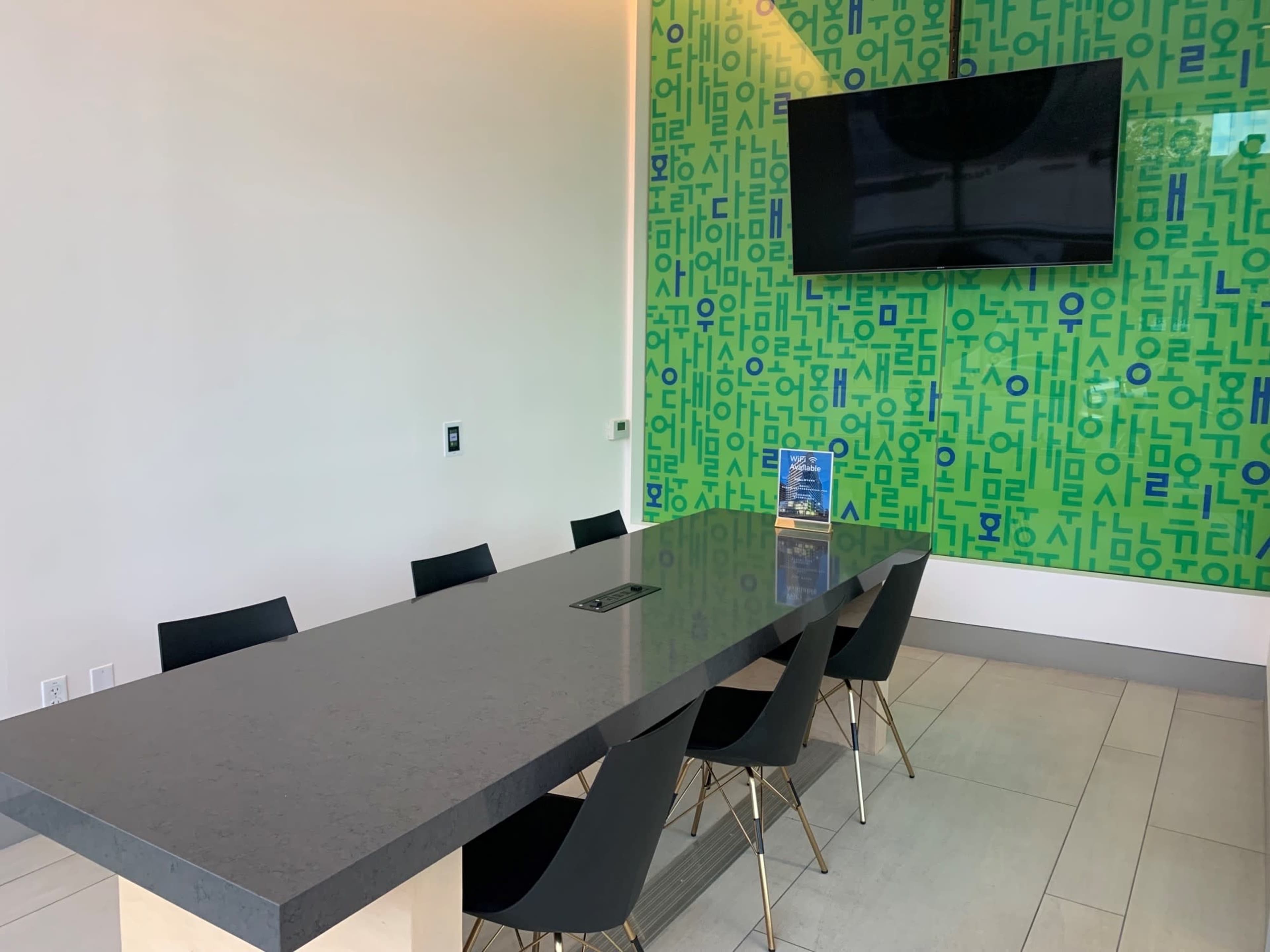 A modern conference room features a long table surrounded by black chairs, with a large television and a green patterned wall in the background.